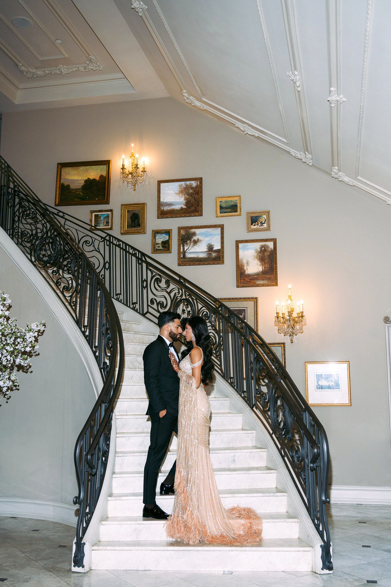 Elegant wedding couple poses on grand white marble staircase with ornate black railing and vintage artwork on walls.
