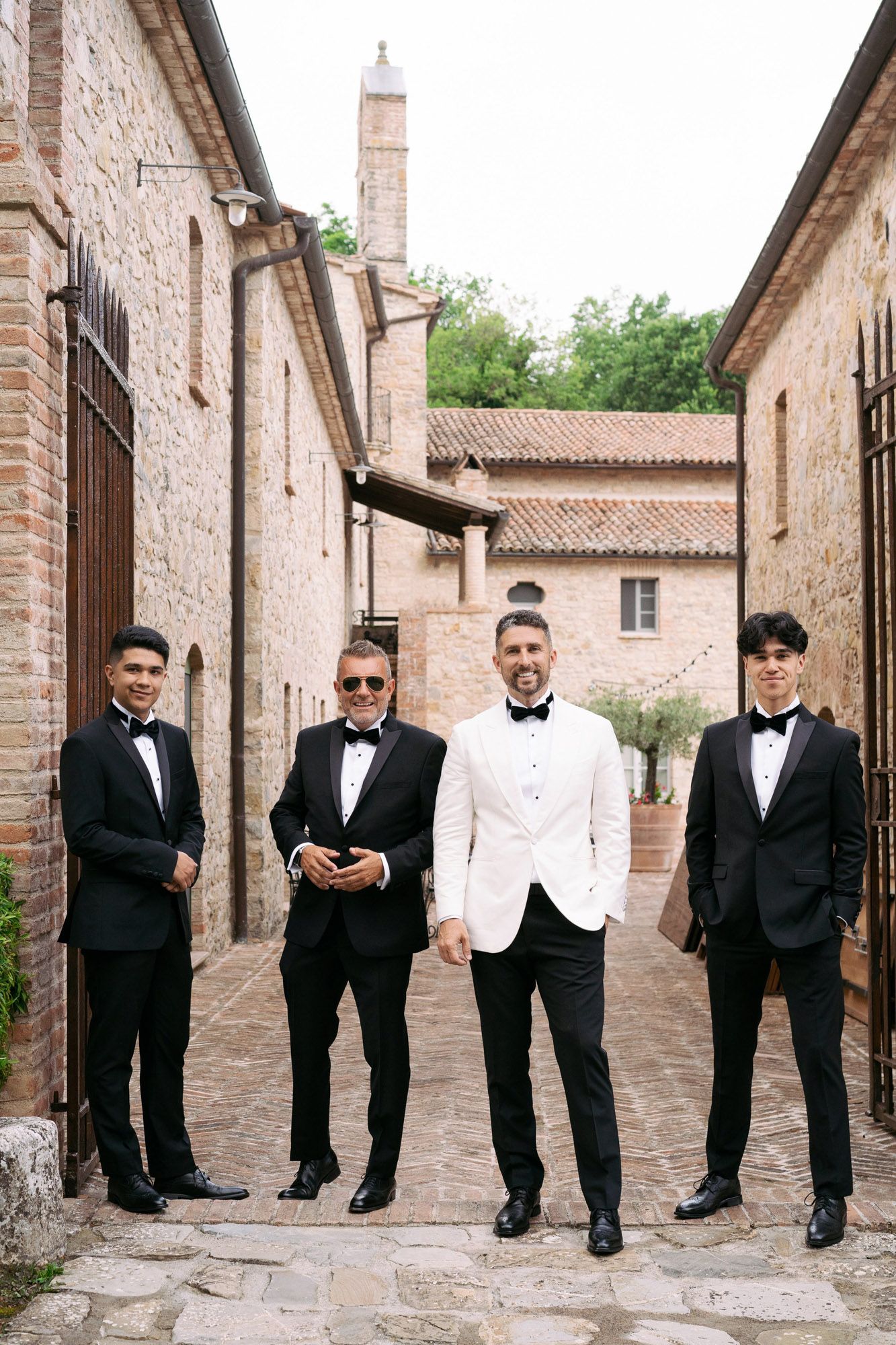 Groomsmen in black and white tuxedos pose together in a rustic stone courtyard setting.