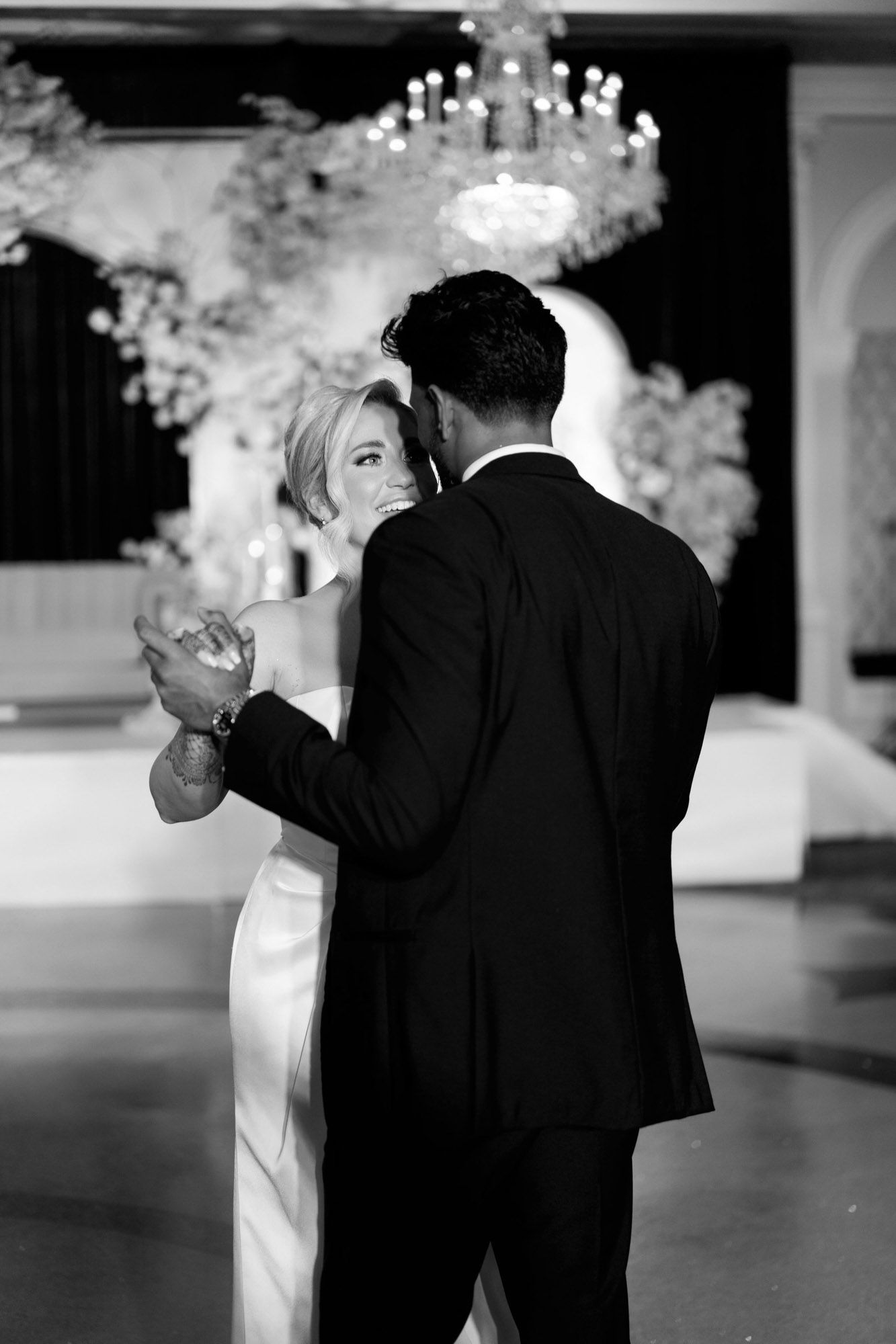 A couple shares their first dance at an elegant wedding reception under a crystal chandelier in black and white photography.