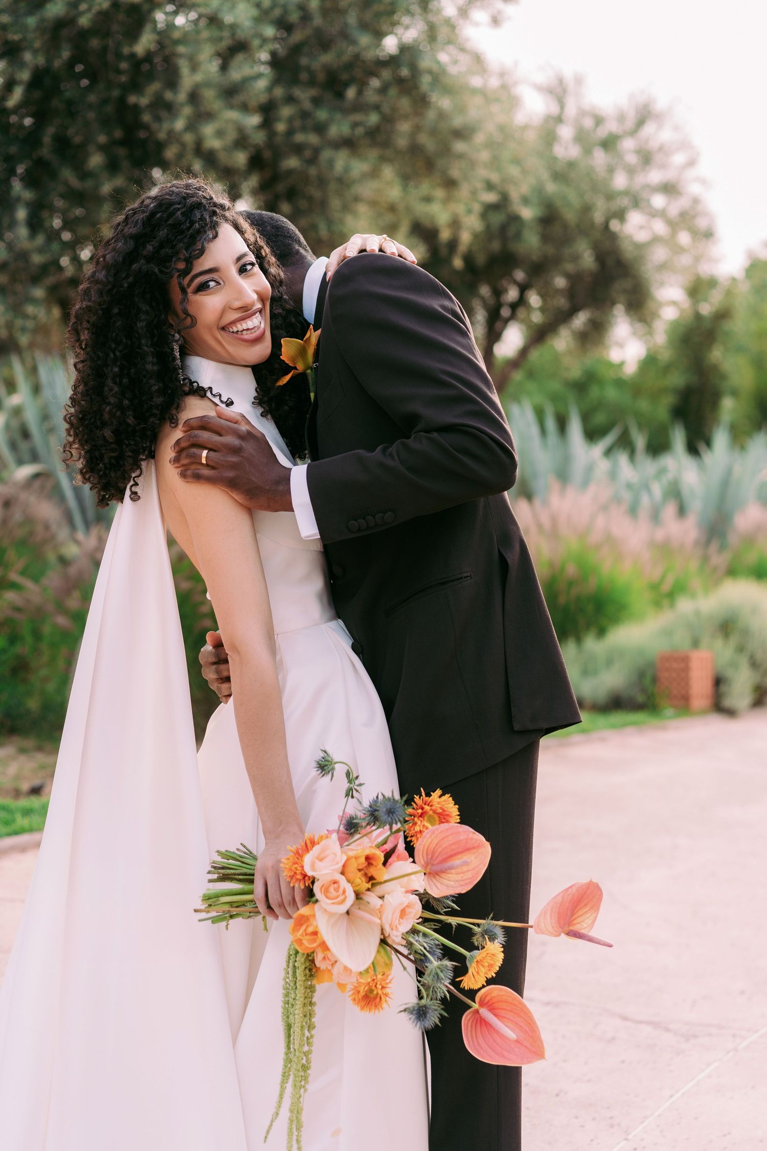 Close-up of wedding couple holding vibrant orange and pink floral bouquet outdoors.