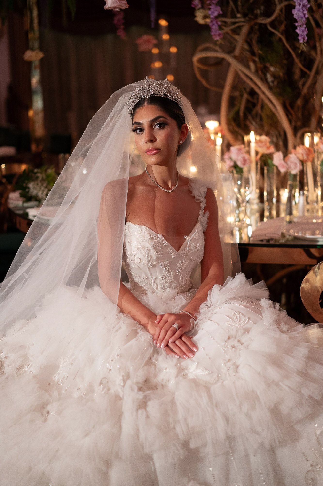 A bride in an elegant white ballgown with ruffled tulle skirt and veil poses in a romantic candlelit setting with hanging wisteria.
