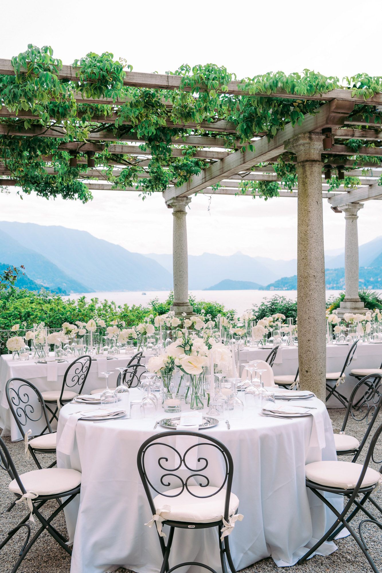 Elegant outdoor wedding reception setup with white tablecloths and wrought iron chairs under a vine-covered pergola overlooking mountains.