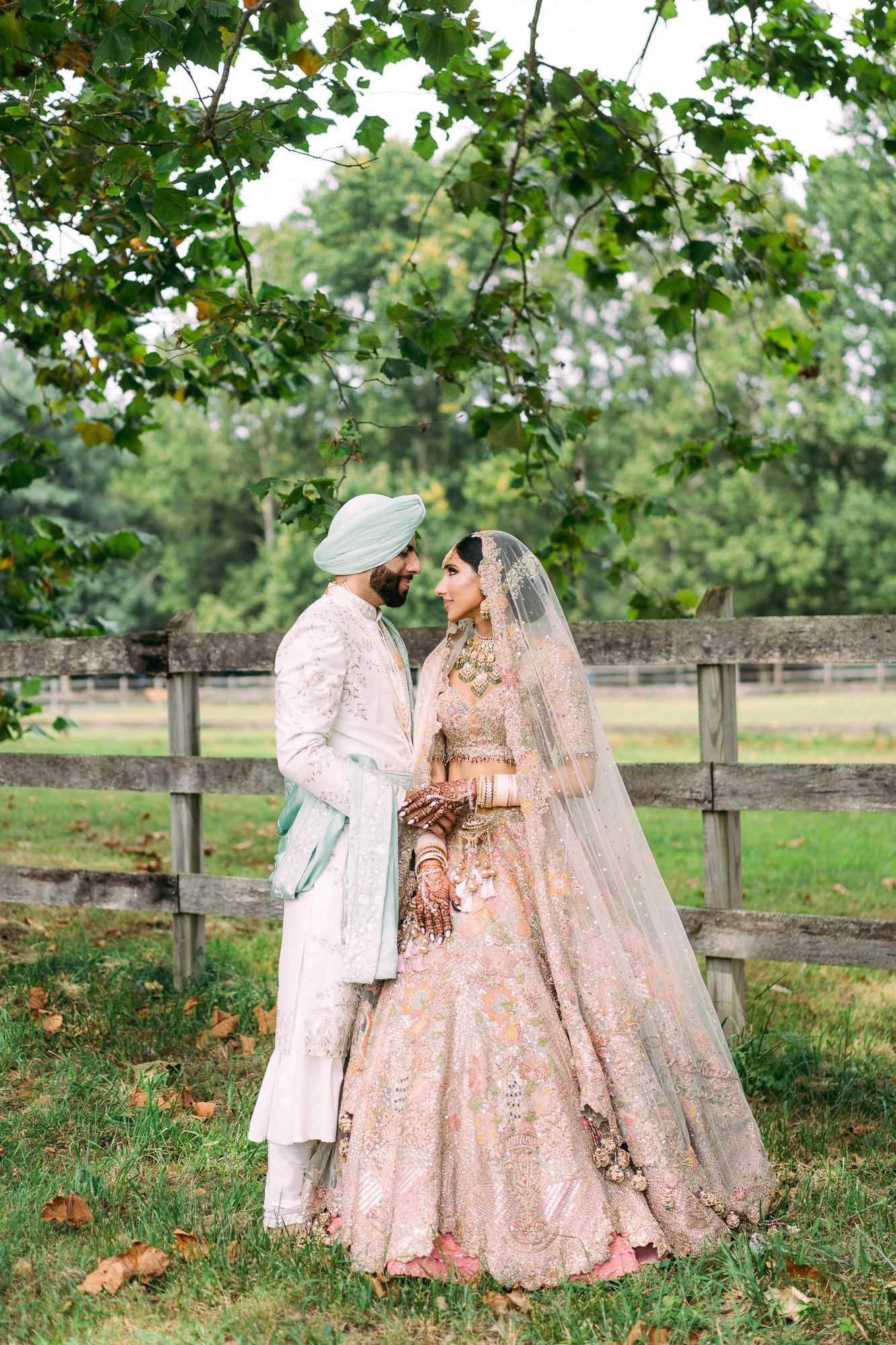 A romantic wedding couple in soft pink traditional Indian attire share an intimate moment in a pastoral garden setting.