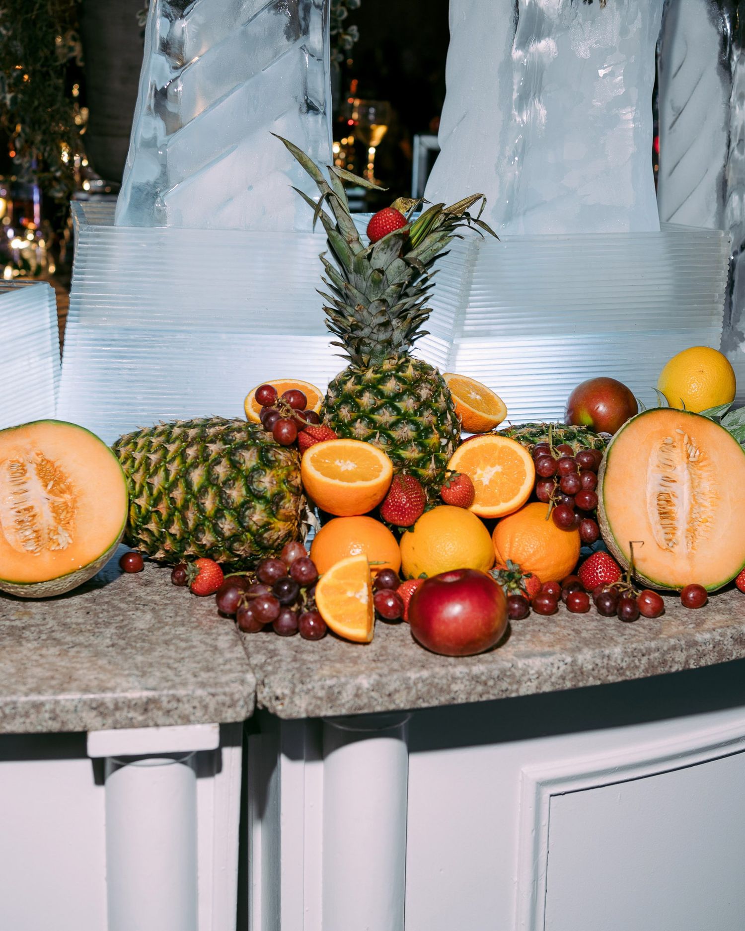Fresh tropical fruit display with pineapples, melons, oranges, grapes and other assorted fruits arranged on white surface.