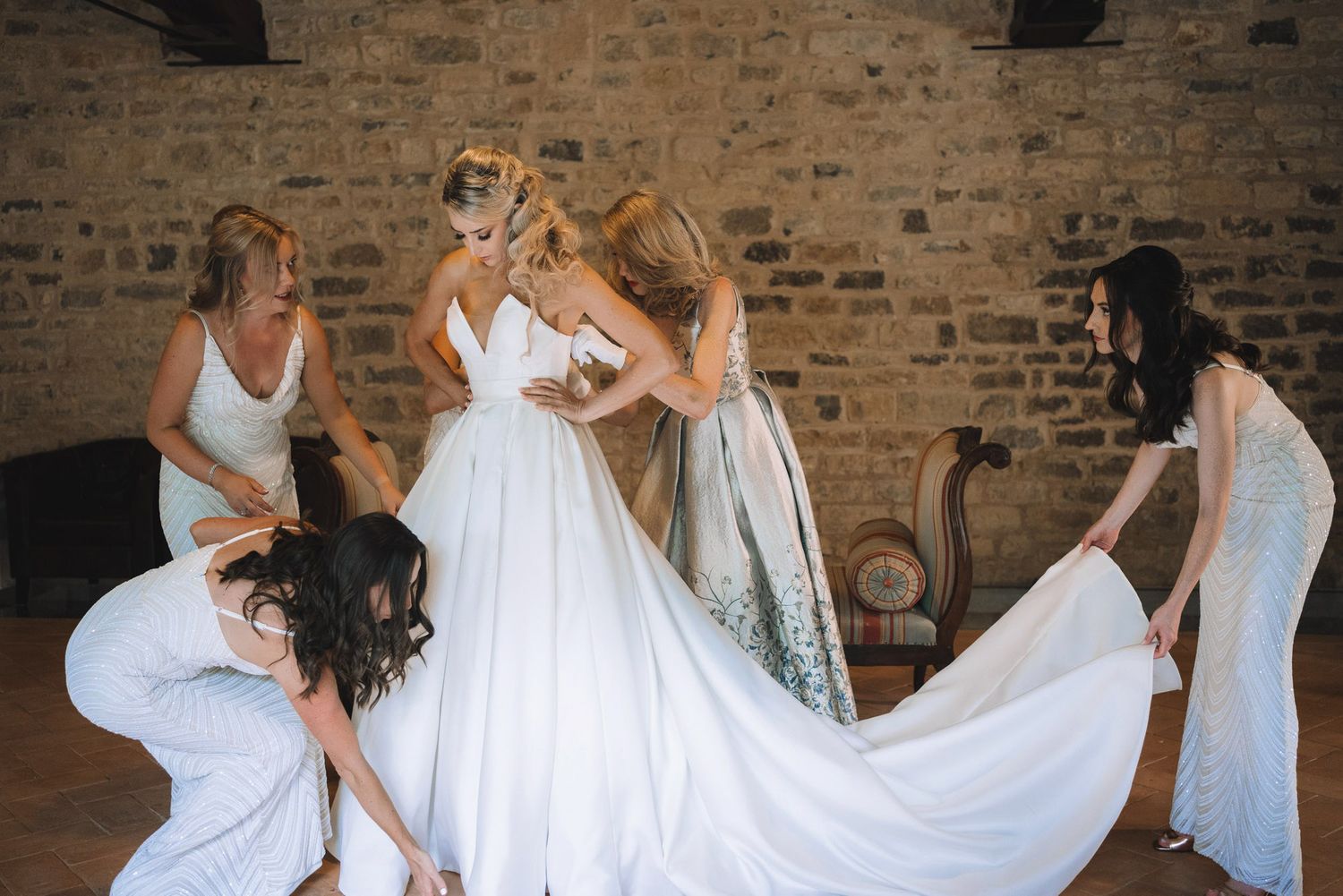 A group in white dresses helps adjust a wedding gown in a rustic brick-walled setting.