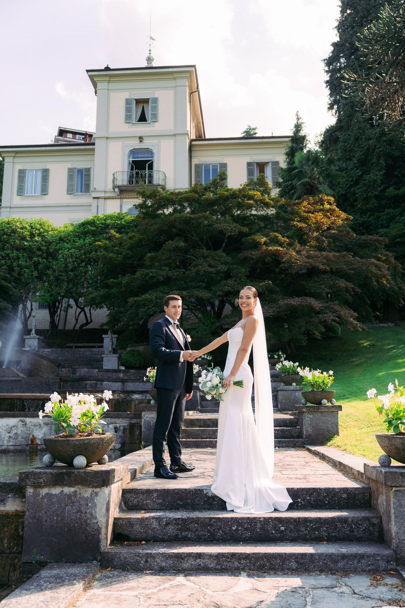 A couple in wedding attire dances playfully on stone garden steps with a stately white villa in the background.
