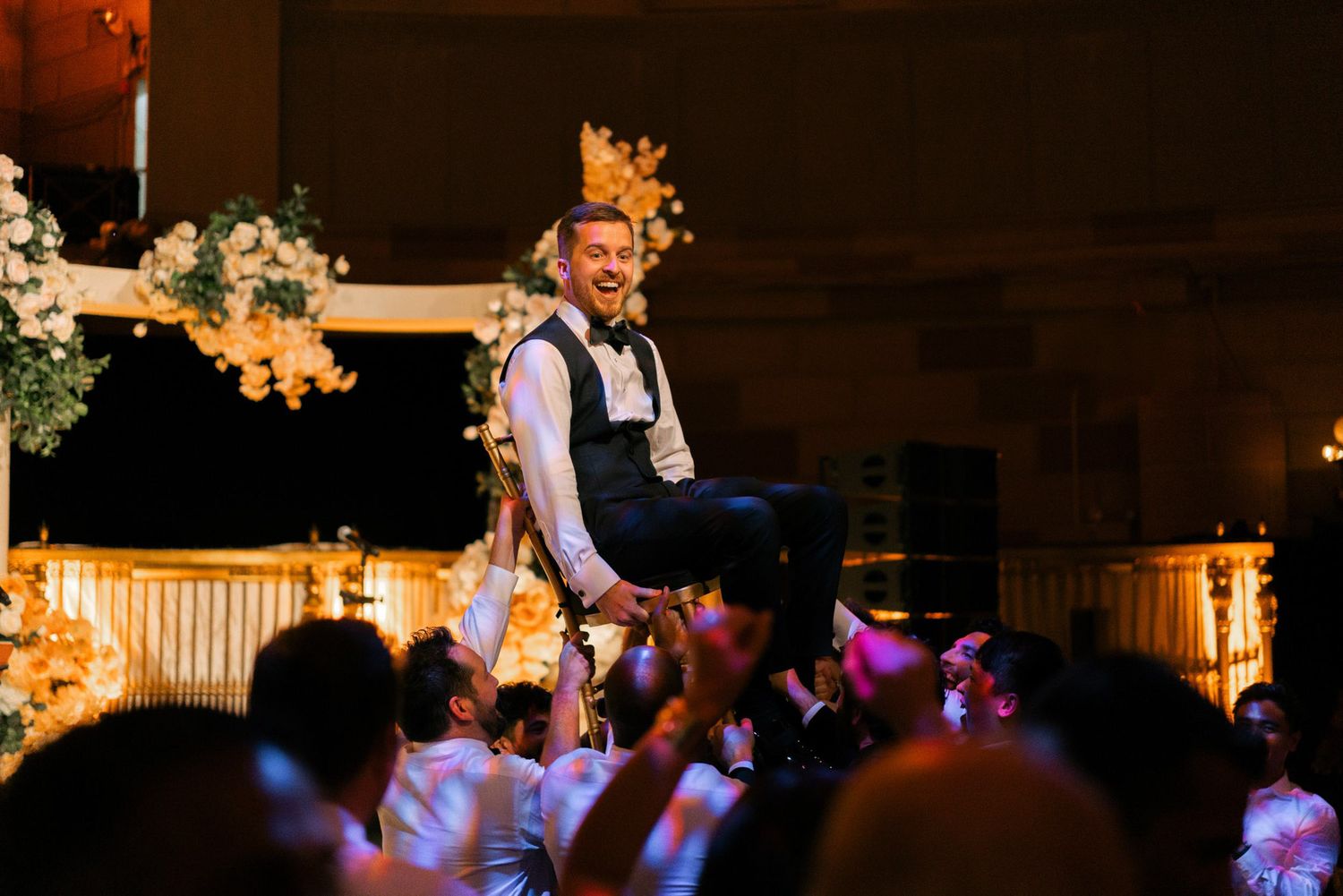 Wedding guests dance and celebrate on a dimly lit dance floor decorated with string lights and floral arrangements.
