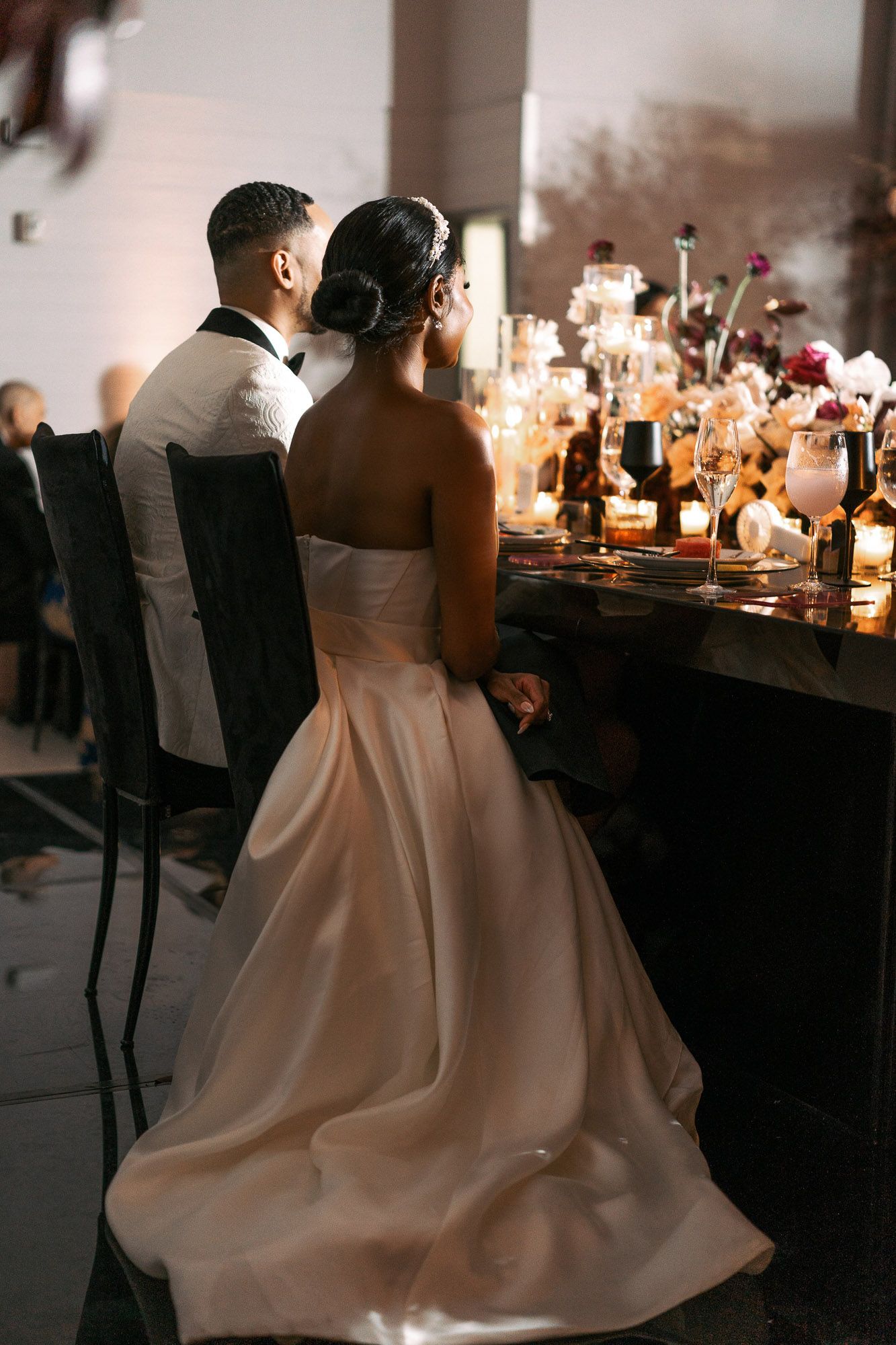 Couple in elegant wedding attire seated at a candlelit reception table with warm ambient lighting.