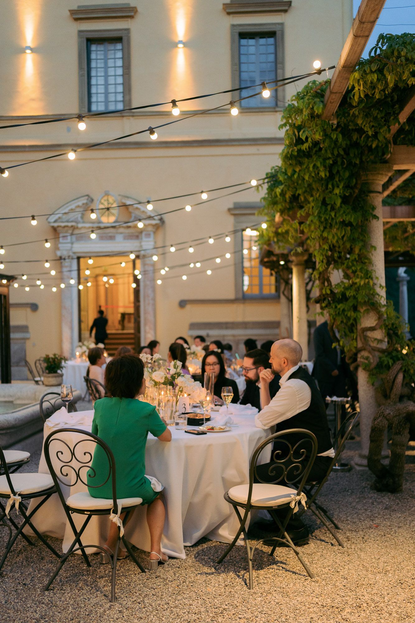 Guests dine at an elegant outdoor patio with string lights and wrought iron chairs at a historic building venue.