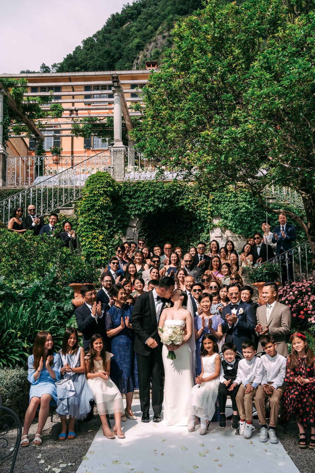 Large group of wedding guests gathered outdoors for a celebratory photo under green trees and brick building.