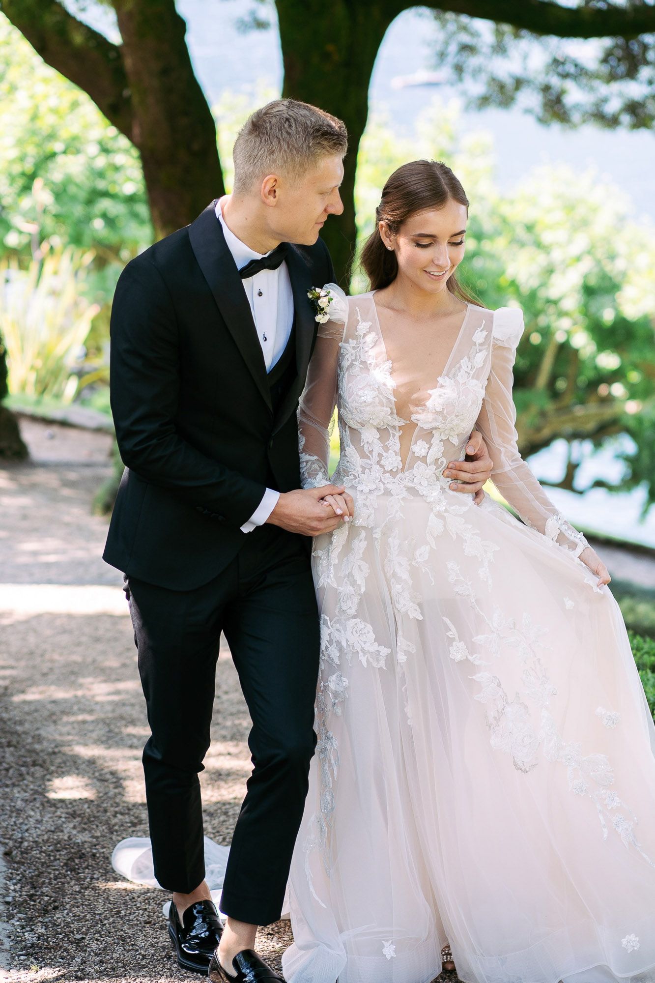 A couple shares a tender moment in wedding attire as the groom helps adjust the bride's dress outdoors.