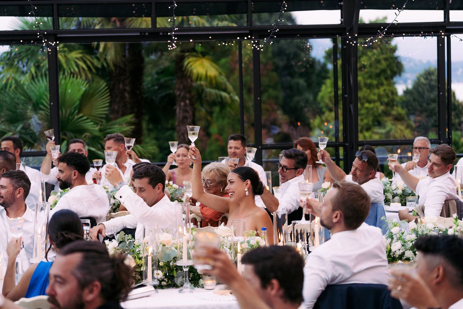 Wedding reception guests raise their glasses in a celebratory toast at an outdoor venue with tropical greenery.