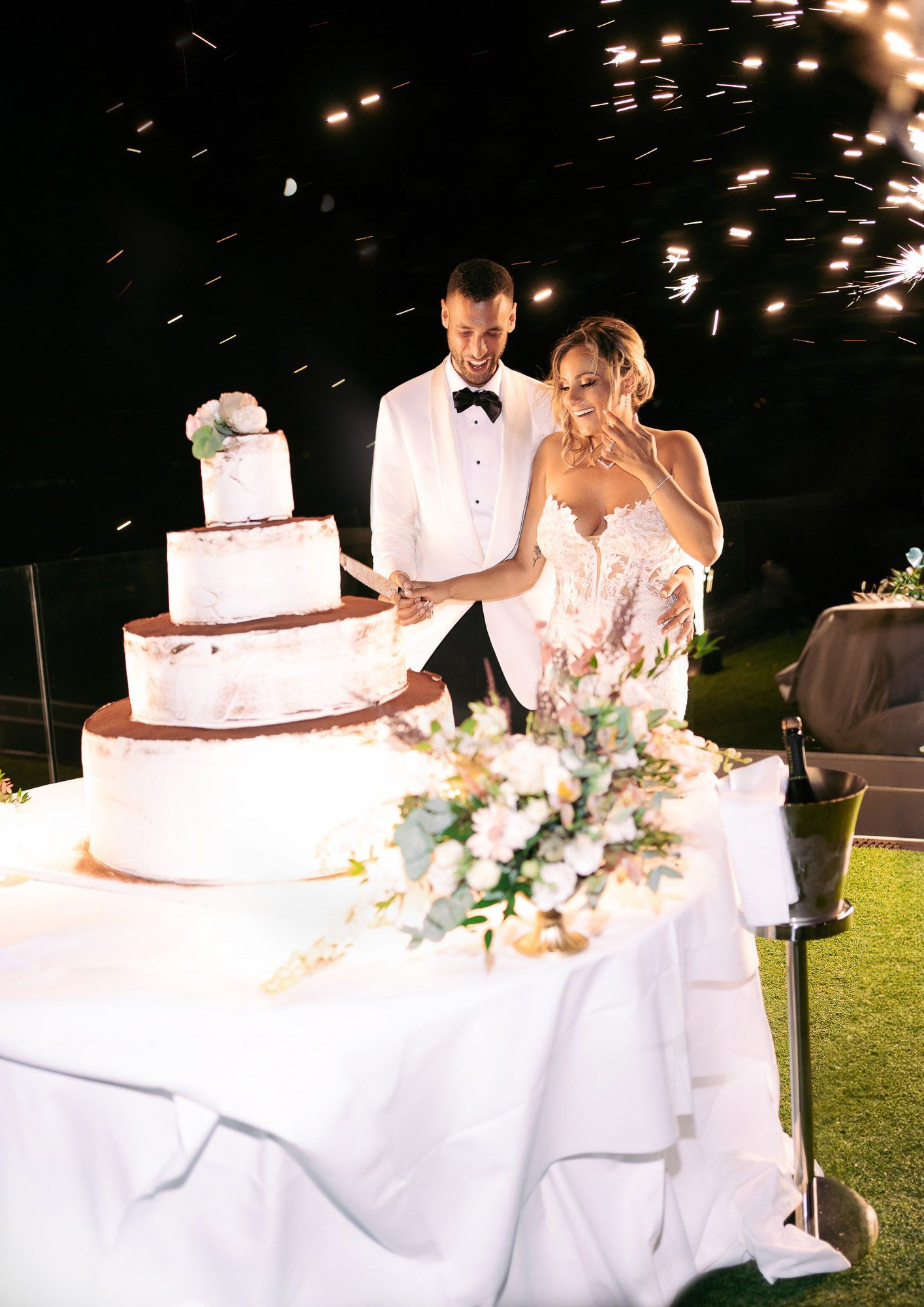Wedding couple cuts their elegant white tiered cake at an outdoor evening reception with sparklers.