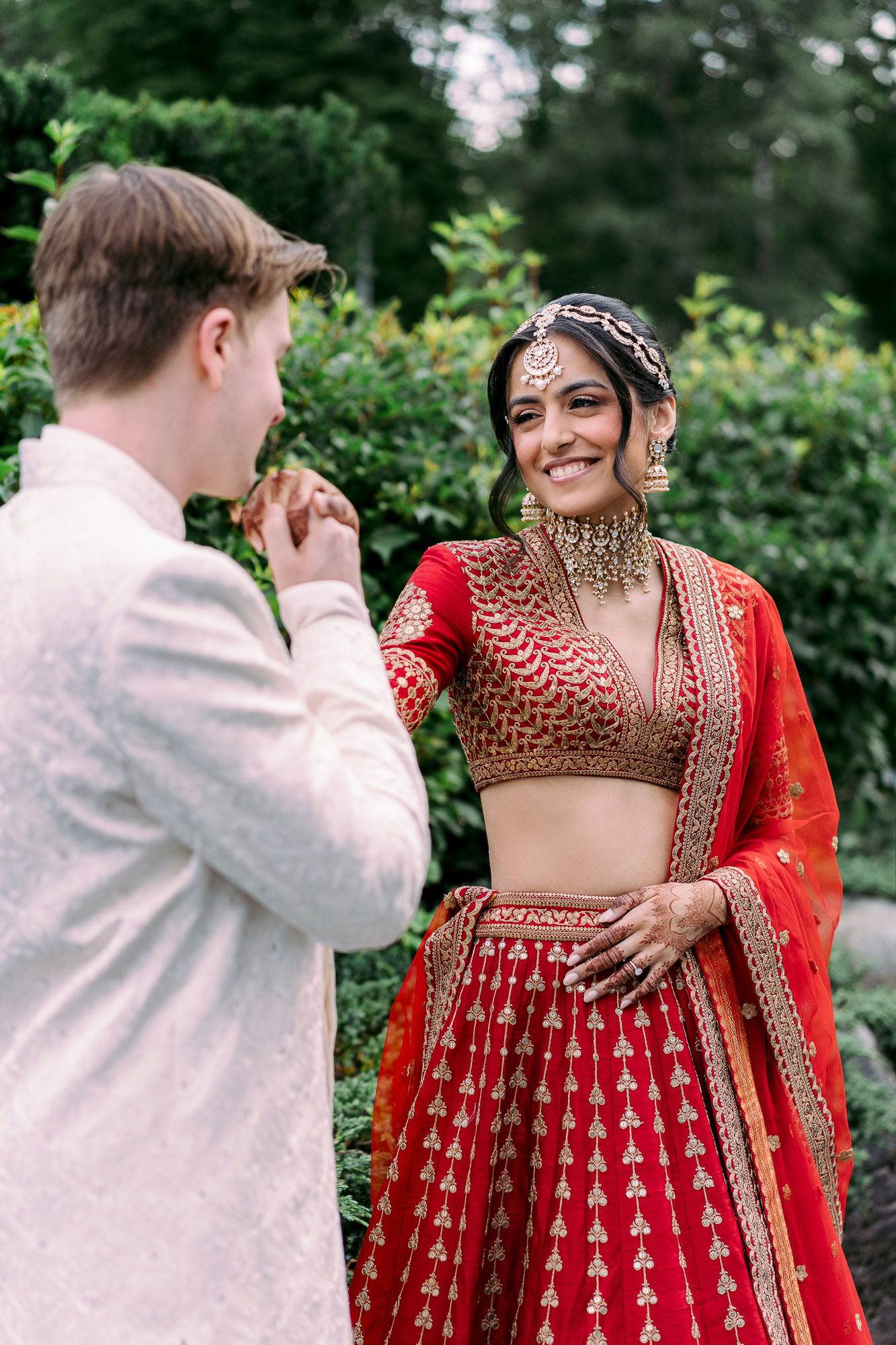 A couple shares tender moments in a garden setting, with the bride wearing a stunning red and gold traditional Indian lehenga.