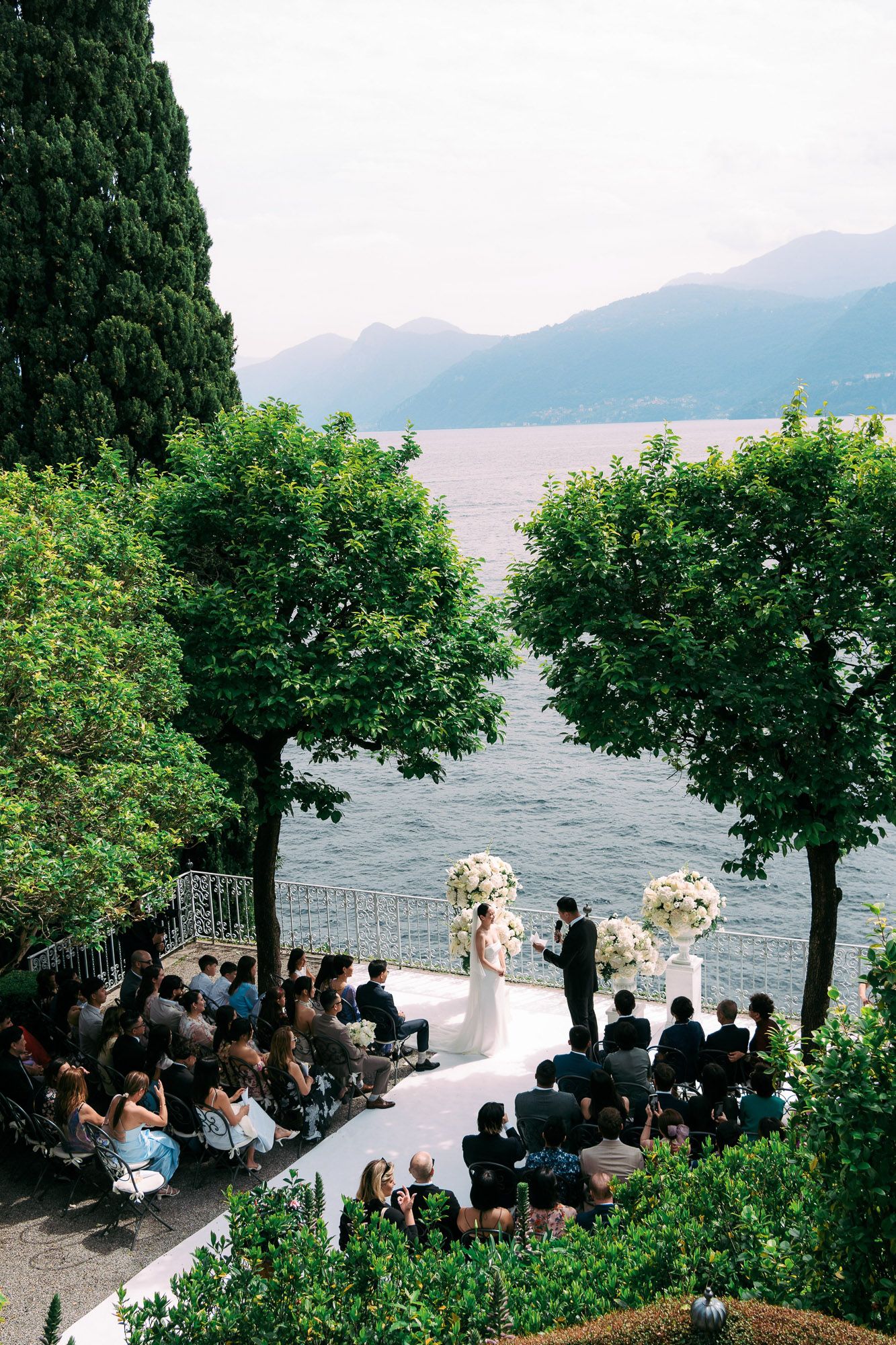 Scenic lakeside wedding ceremony set against mountains with guests seated among lush greenery and trees.