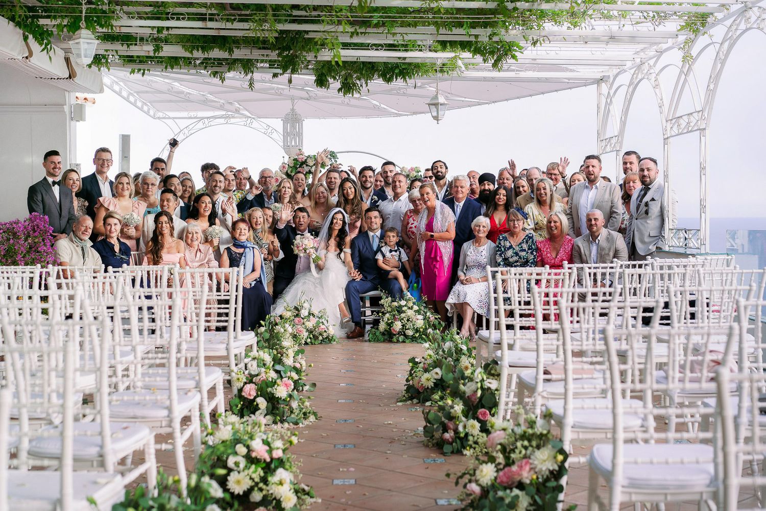Wedding guests gather at an outdoor venue with white chairs and floral decorations lining the aisle for a seaside ceremony.