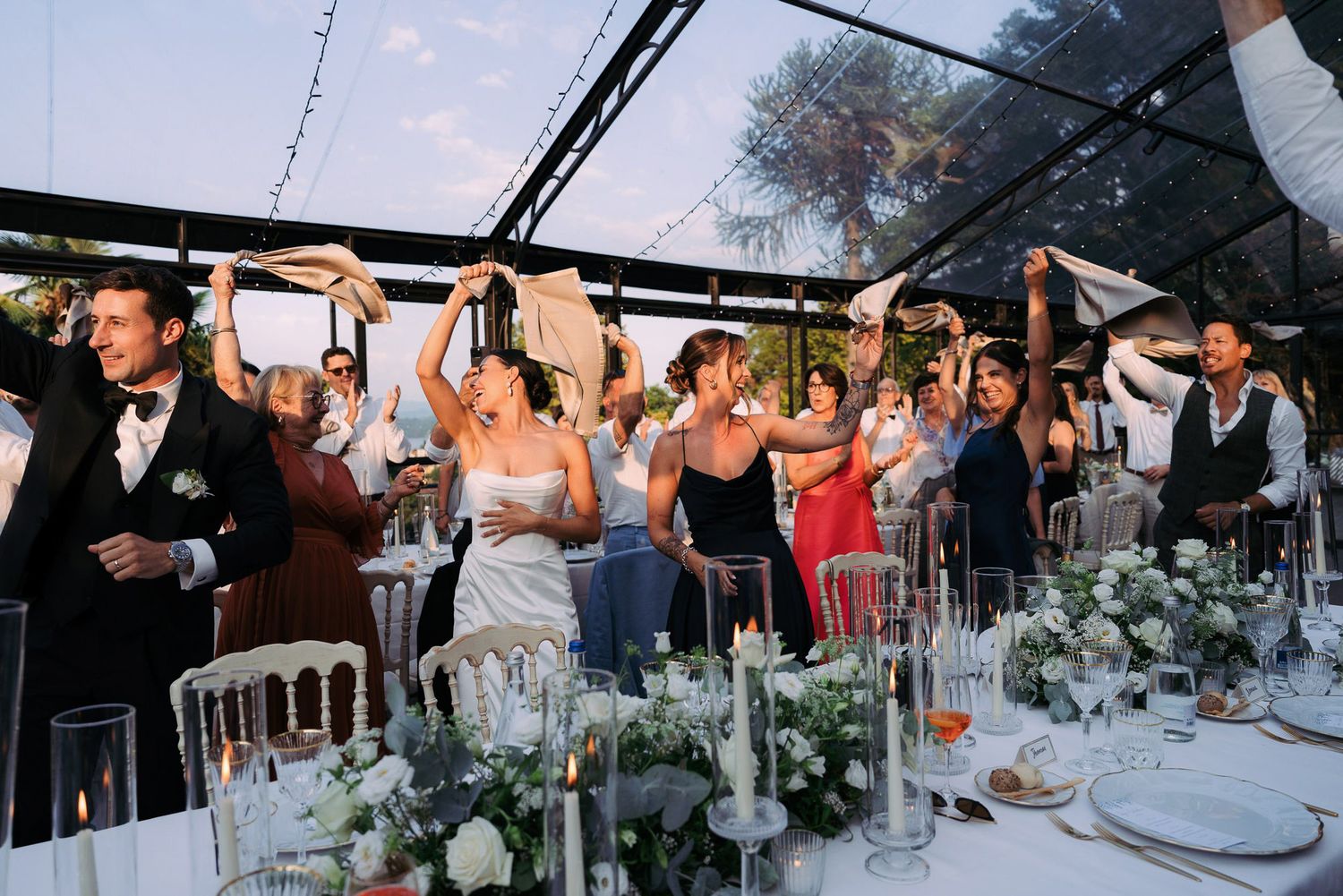 Wedding reception guests celebrate and dance together under an elegant white tent decorated with floral arrangements.
