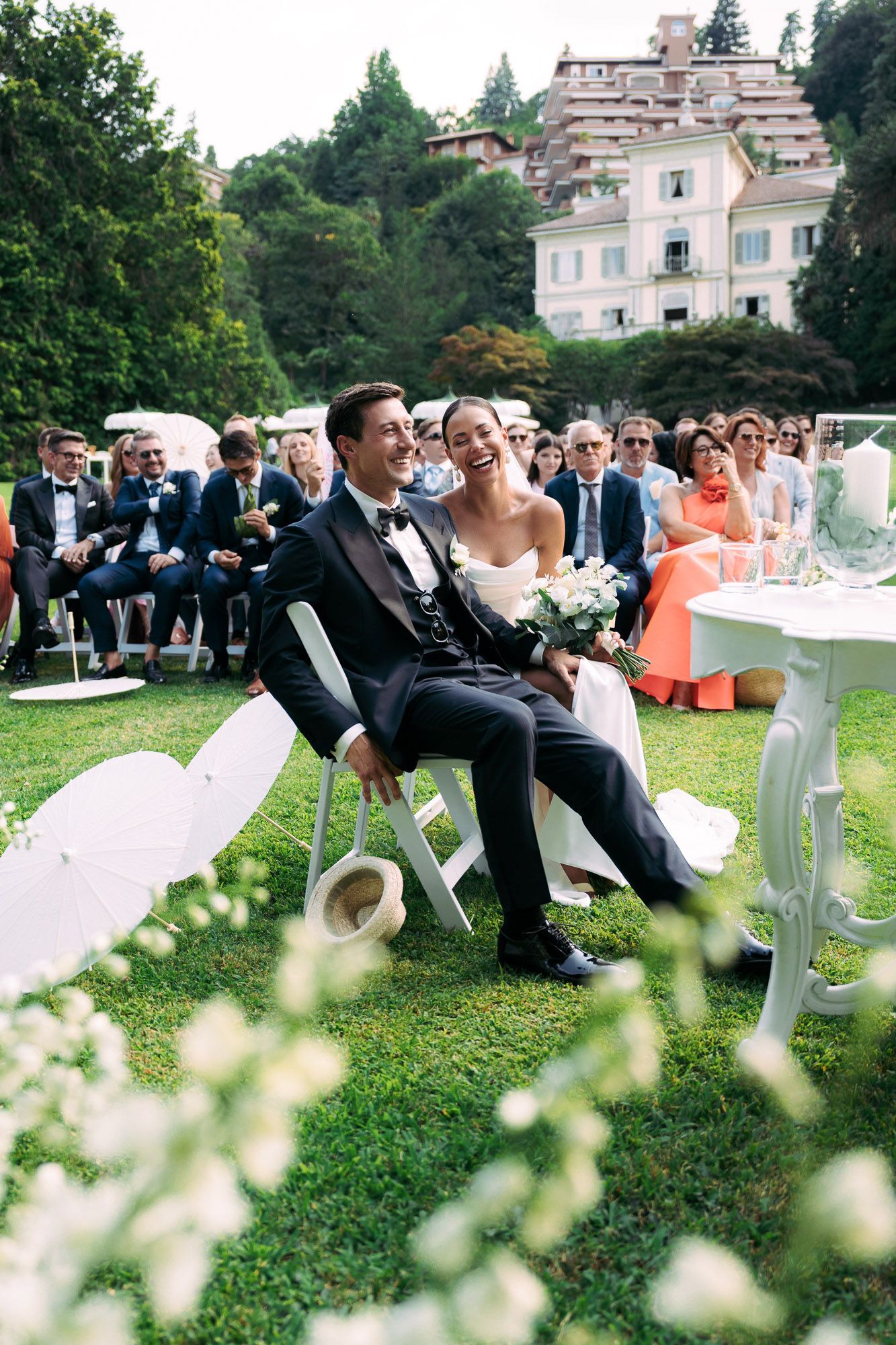 Wedding guests relax on white chairs arranged on a grassy lawn with blooming flowers in the foreground at an outdoor ceremony.
