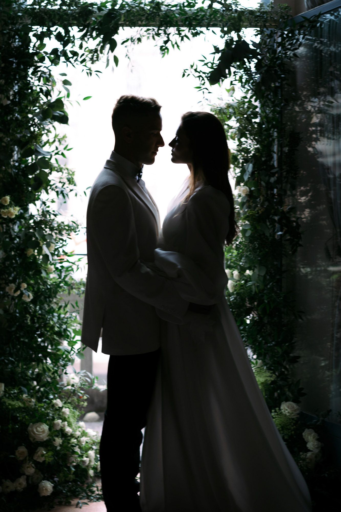 Silhouetted couple in wedding attire share an intimate moment under a lush green archway with natural light behind them.