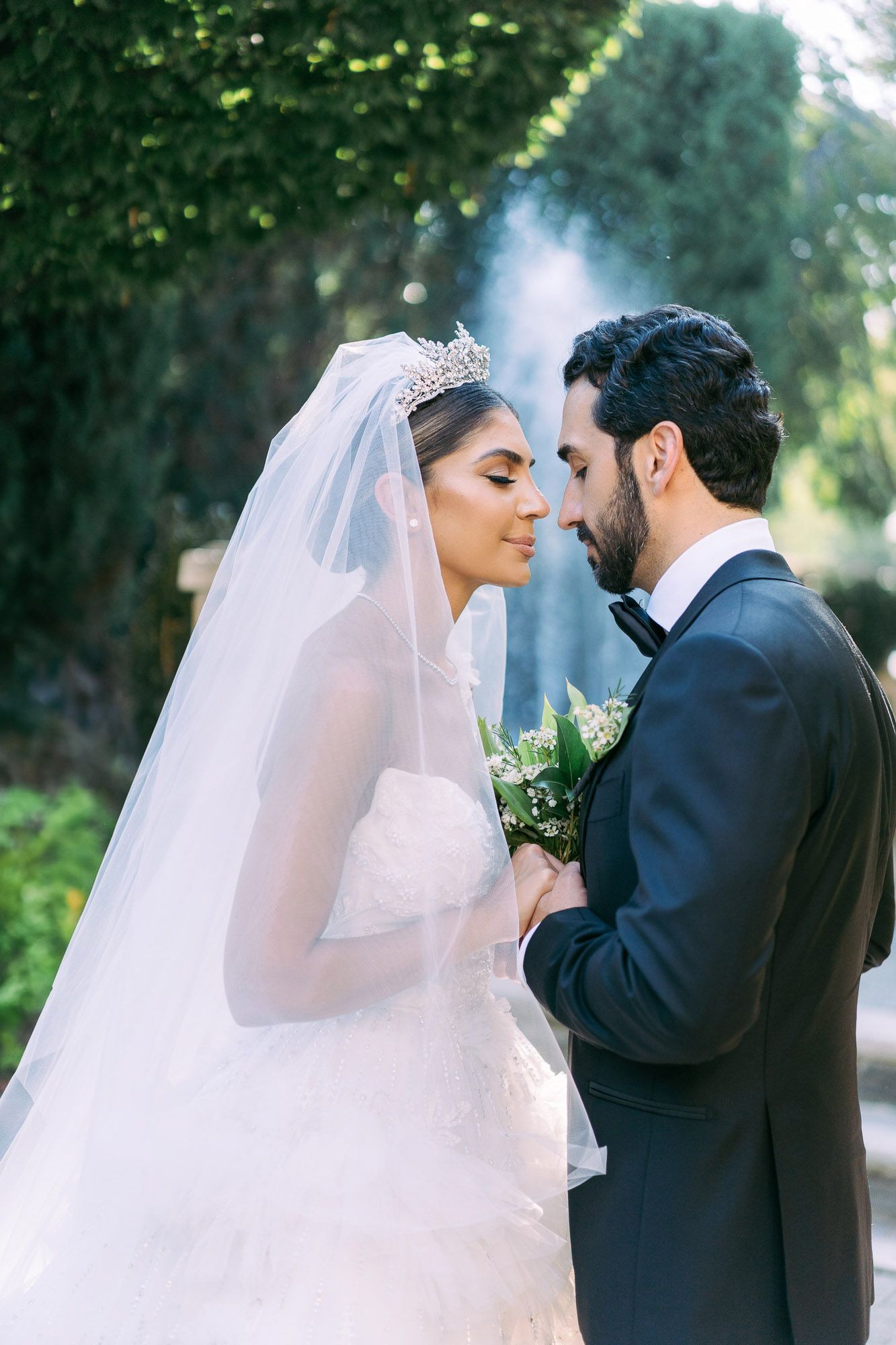 Romantic wedding couple shares intimate moments in a garden setting with flowing white veil and formal attire.