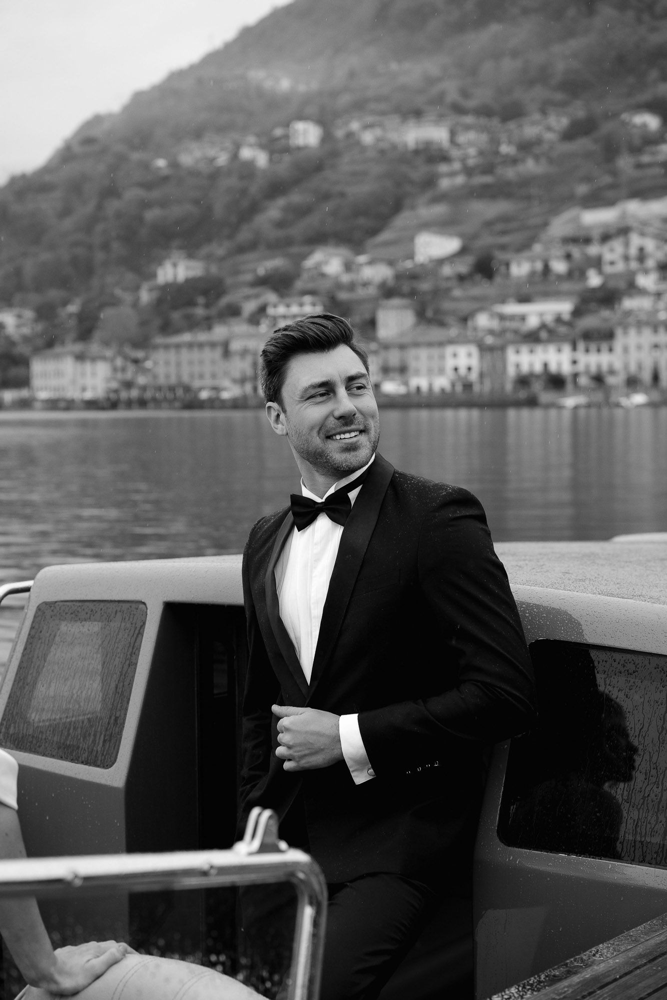 A person in a tuxedo sits in a vintage boat on a scenic lake with mountains and buildings in the background.