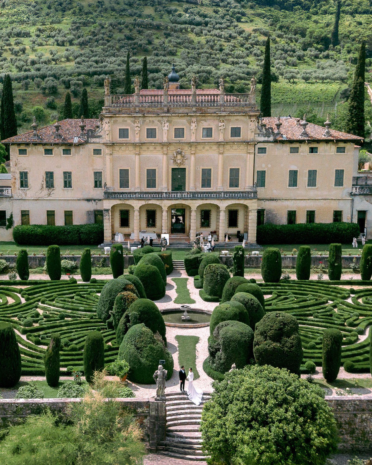 Aerial view of a grand Italian villa with meticulously manicured formal gardens featuring ornate hedges and geometric patterns.