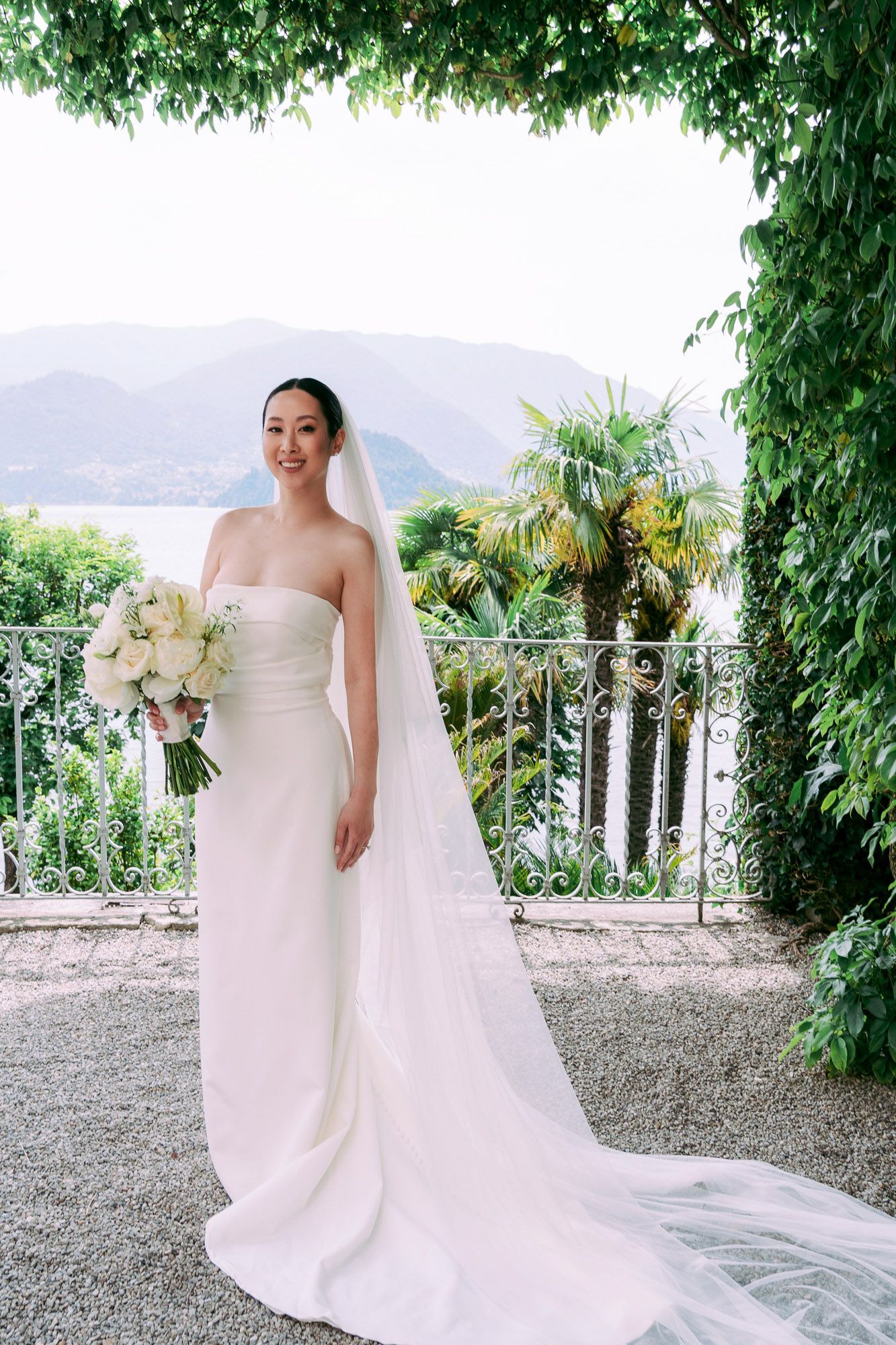 A bride in an elegant white wedding dress with a flowing veil poses on a terrace overlooking a scenic mountain landscape.