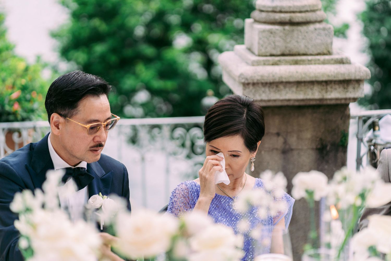 A couple shares a tender moment in a garden setting with stone pillars and purple flowers in the background.