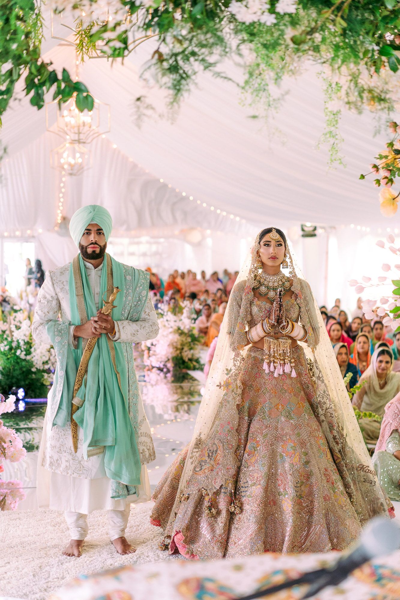 Traditional Indian wedding couple in mint green and gold embellished outfits standing together at decorated venue.