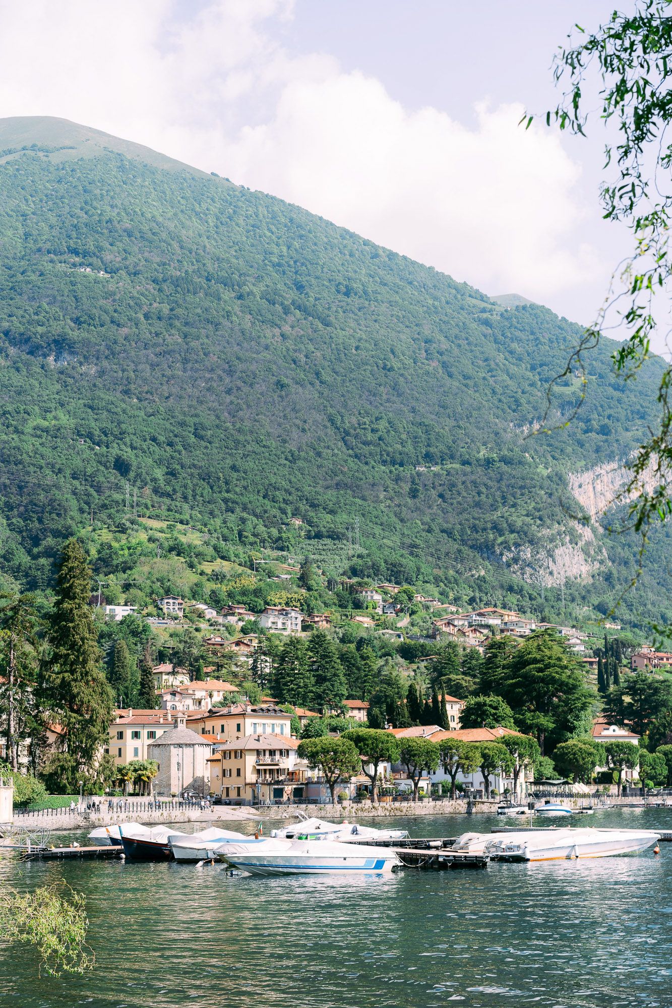 Scenic lakeside view of Italian village with boats docked along Como Lake shore against mountainous backdrop.