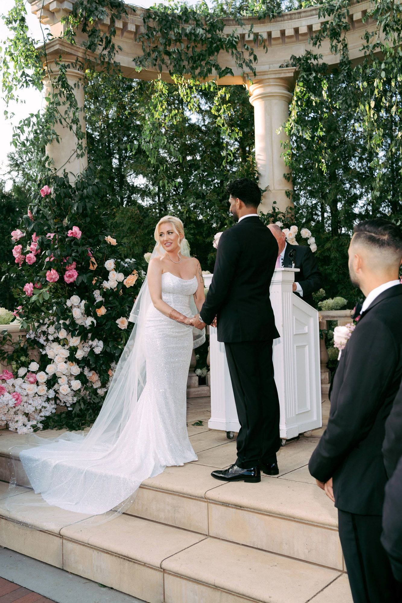 A wedding couple shares a tender moment on stone steps surrounded by climbing roses and greenery at an elegant venue.
