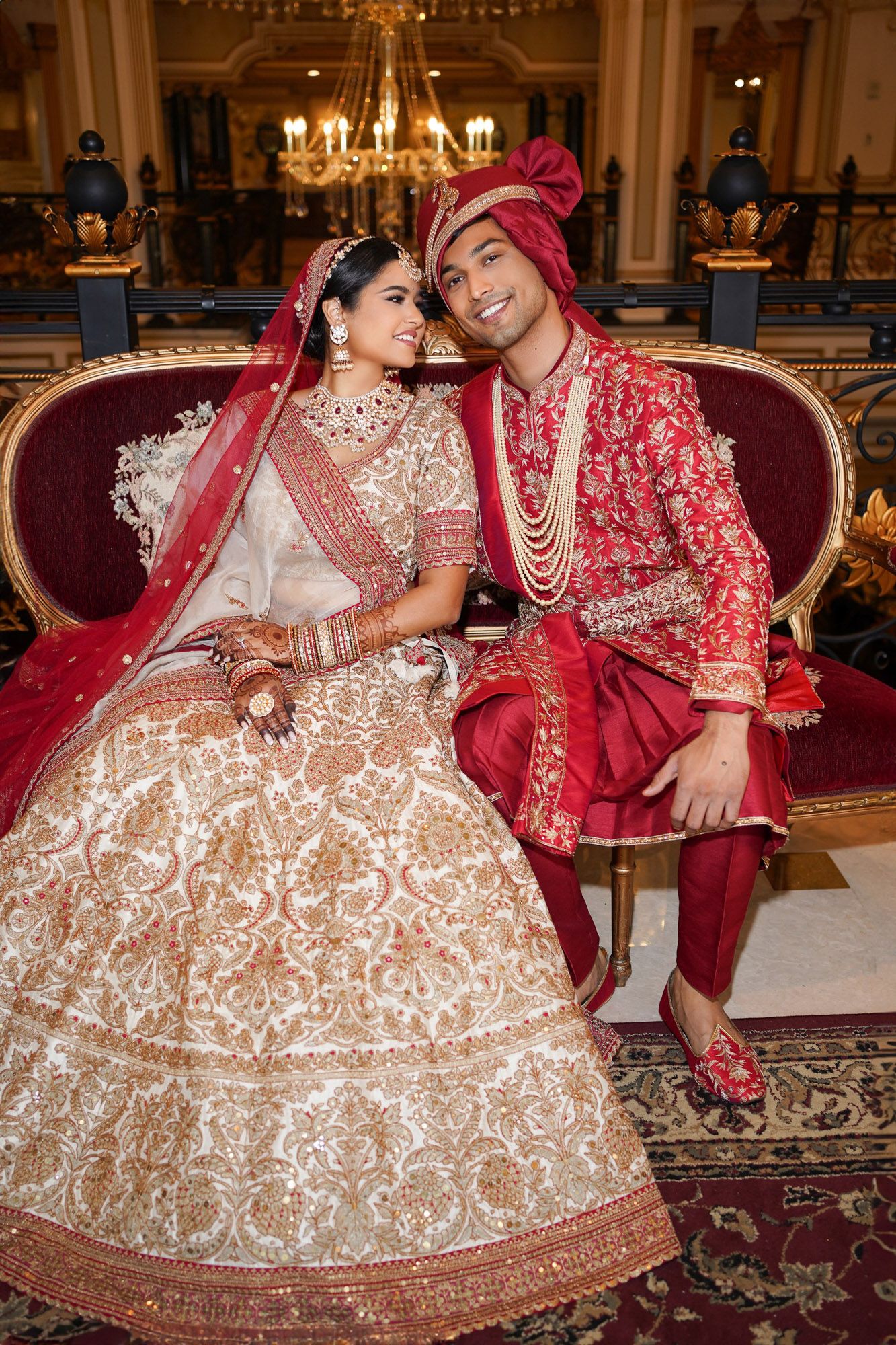 Couple in traditional Indian wedding attire with red and cream lehenga posing on ornate furniture in elegant venue.