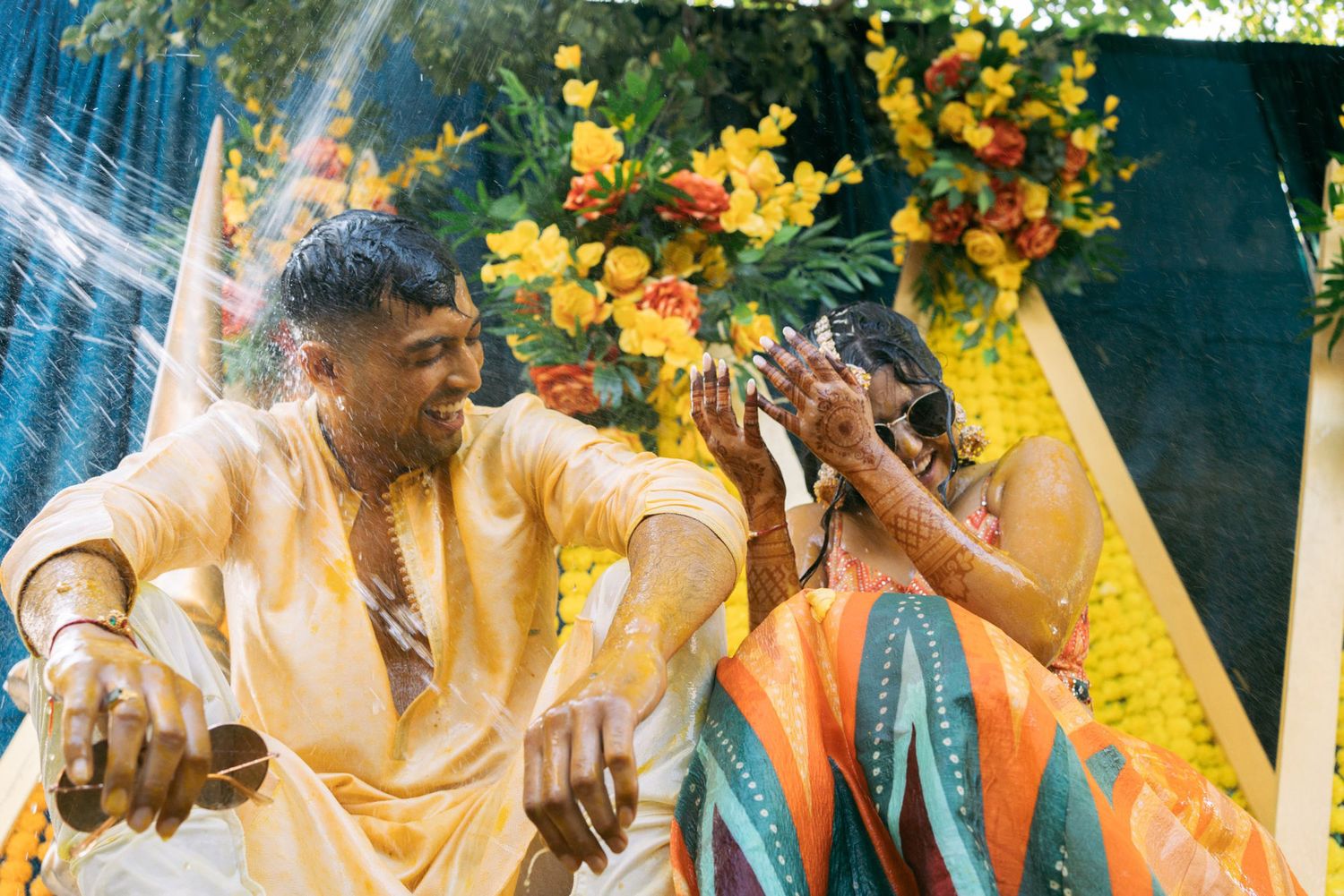 Joyful celebration with turmeric powder during traditional Haldi ceremony with colorful floral decorations in background.