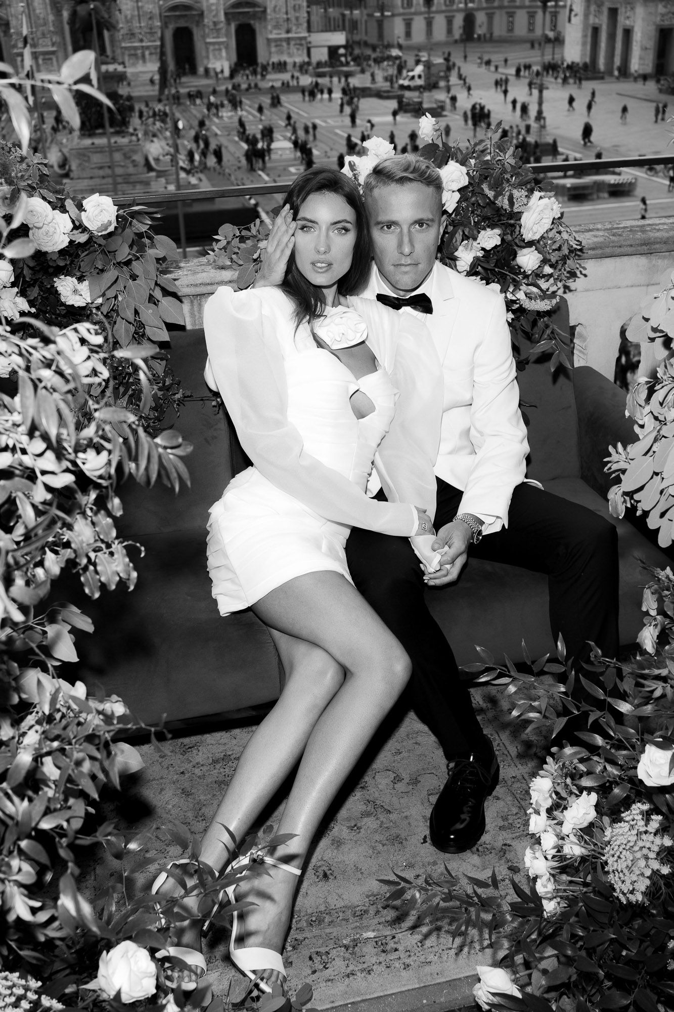 A couple in white formal attire poses together surrounded by floral decorations in a black and white photograph.