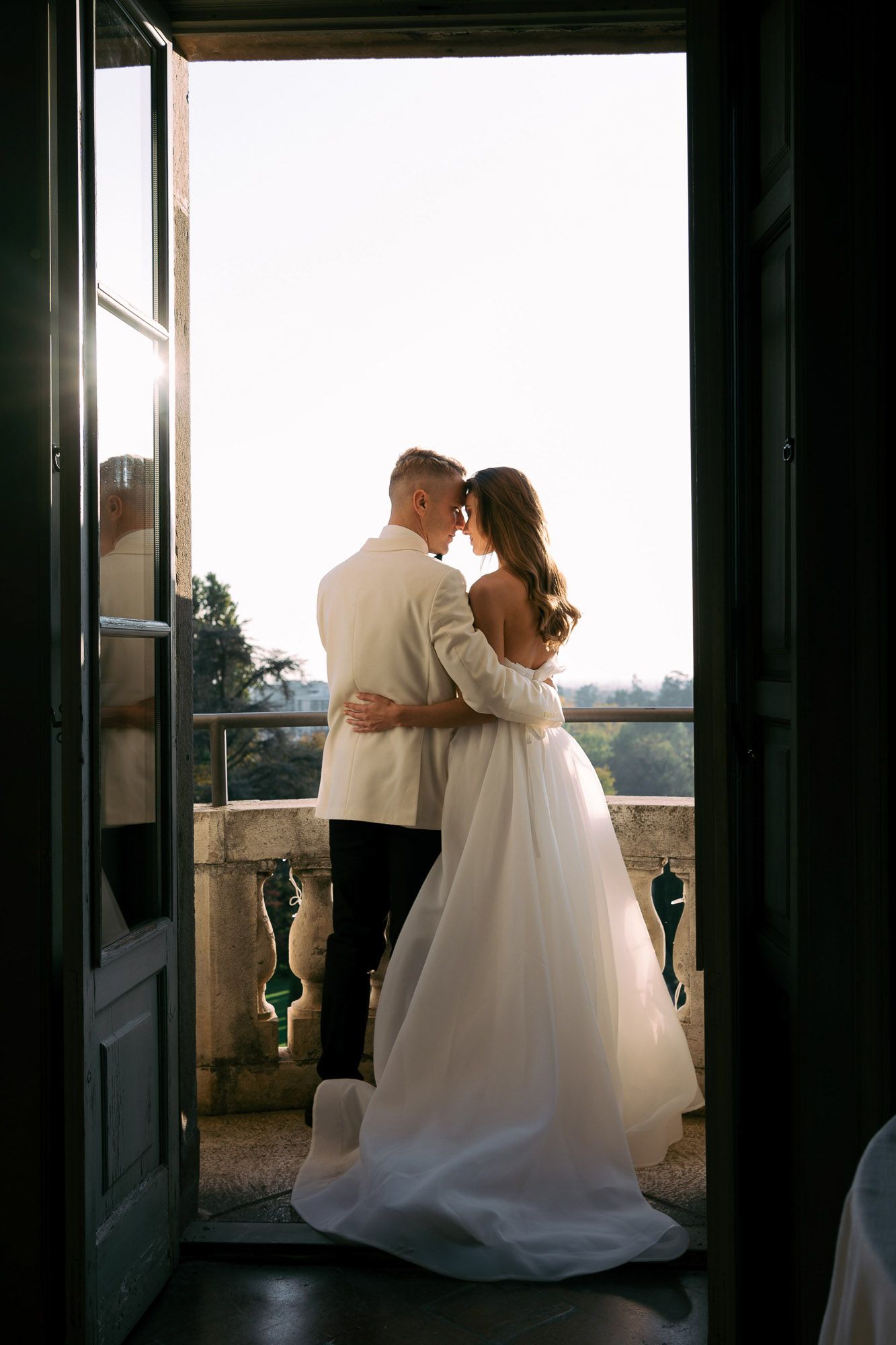 A romantic silhouette of a couple in wedding attire embracing by a window during sunset.