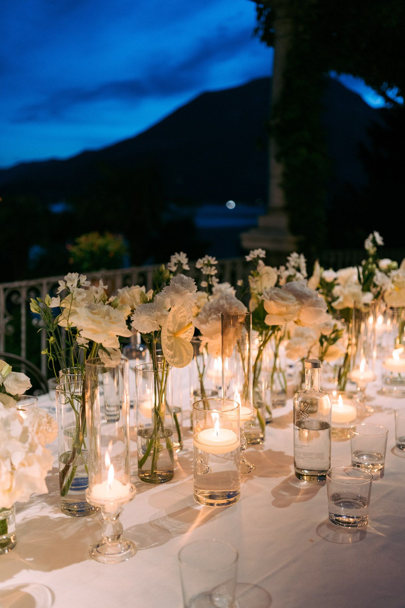Elegant table setting with glowing candles and white flowers in glass vases against a twilight mountain backdrop.