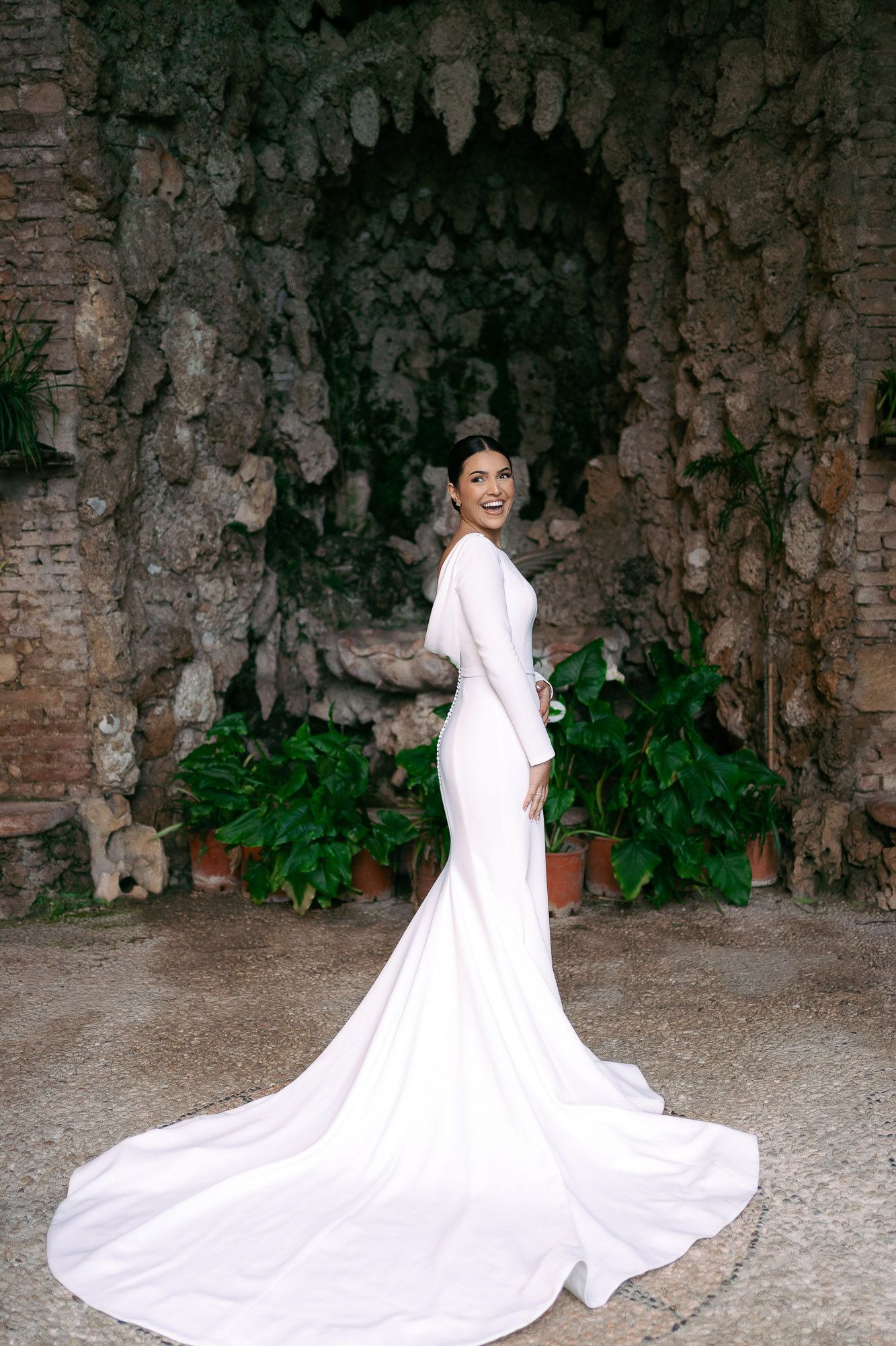 A bride in a flowing white gown with a dramatic train poses against a rustic stone wall backdrop.
