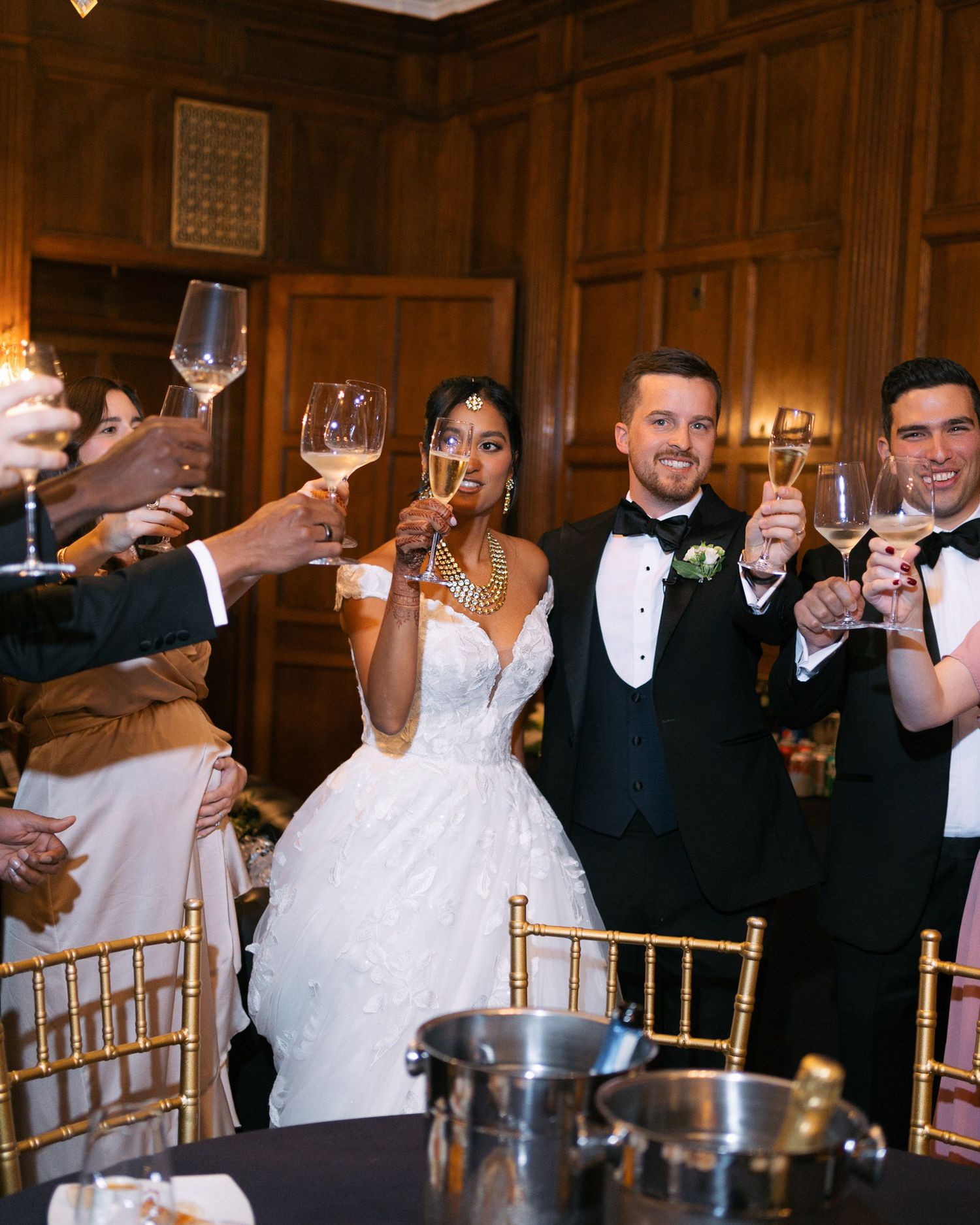 Wedding guests raise glasses in a toast during an elegant reception in a wood-paneled ballroom.