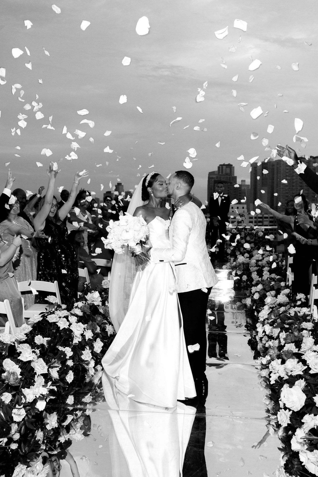 A romantic wedding ceremony moment captured in black and white as flower petals fall from above during the celebration.