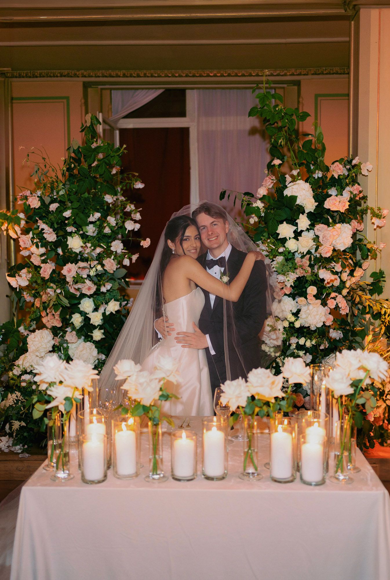 Elegant wedding reception table decorated with glowing candles and lush floral arrangements in white and peach tones.