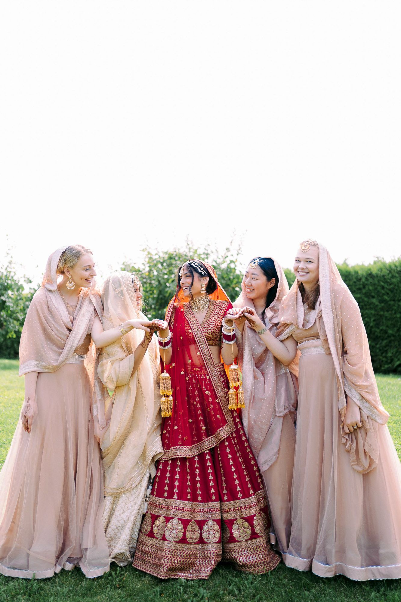 Bridesmaids in blush pink outfits share a joyful moment with bride wearing traditional red Indian lehenga in garden setting.