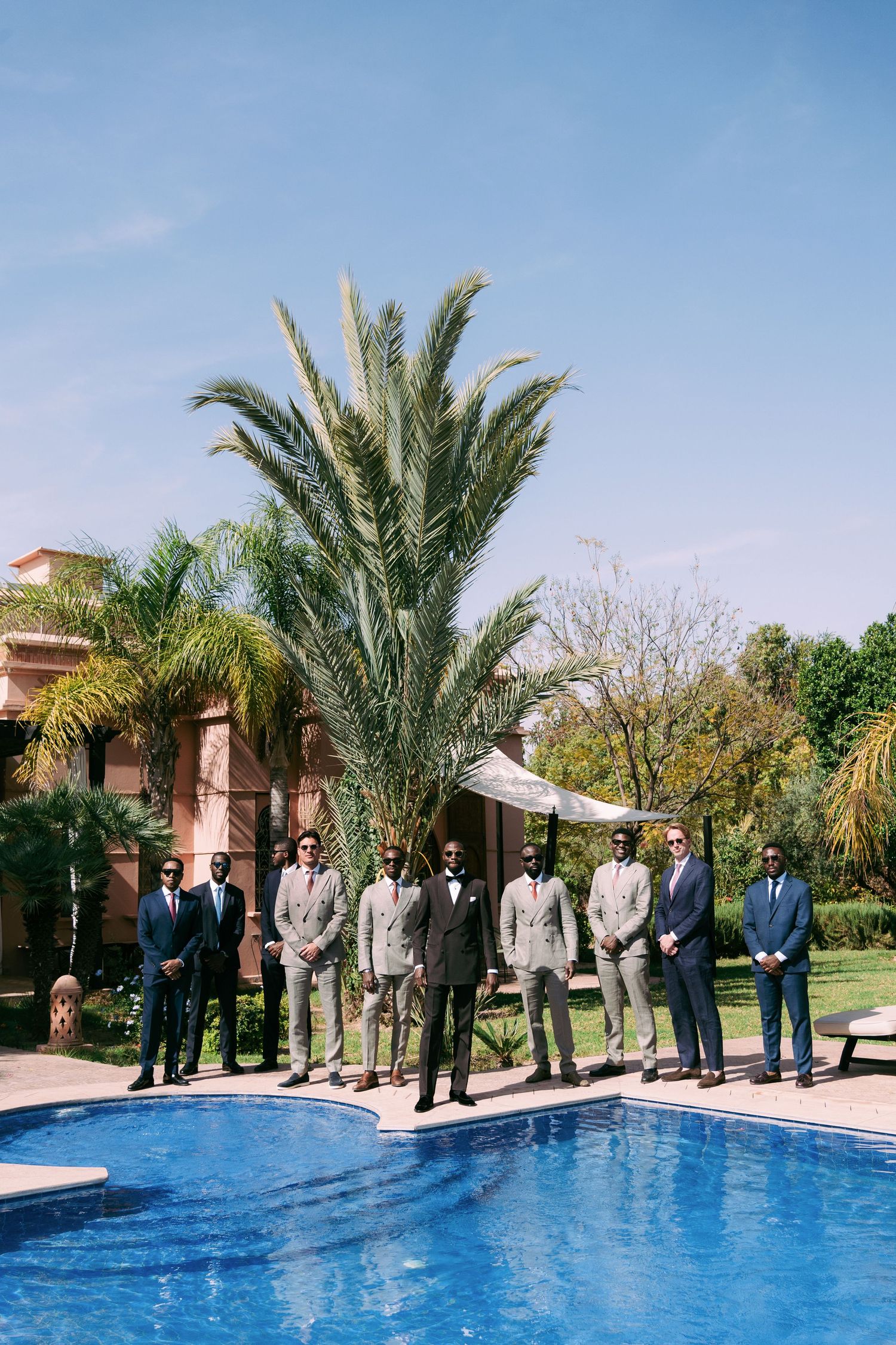 A lineup of formally dressed wedding party members posing by a swimming pool at a luxury venue.