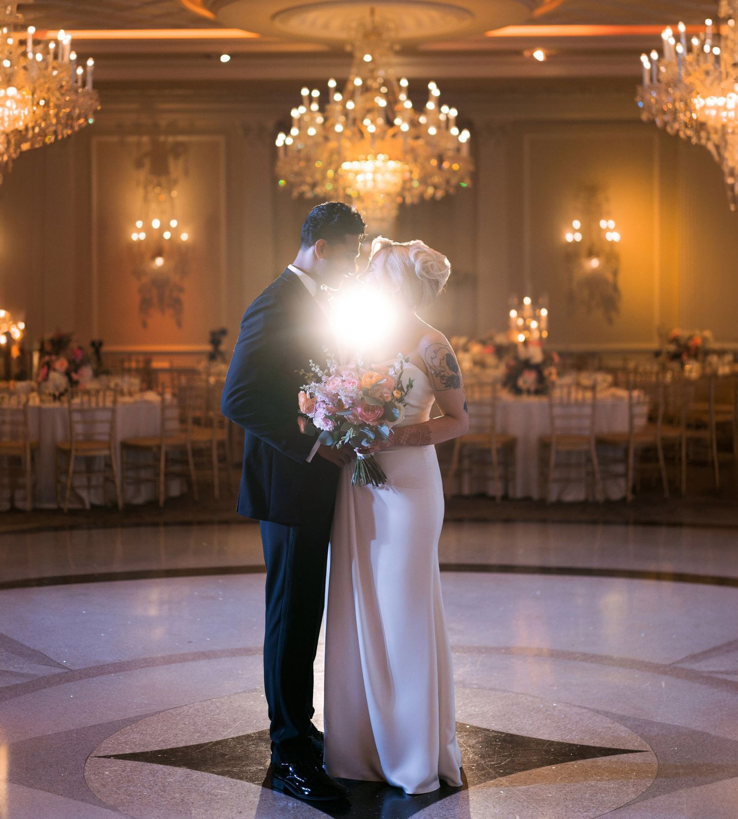 Elegant wedding couple dancing in a luxurious ballroom with crystal chandeliers and warm lighting.