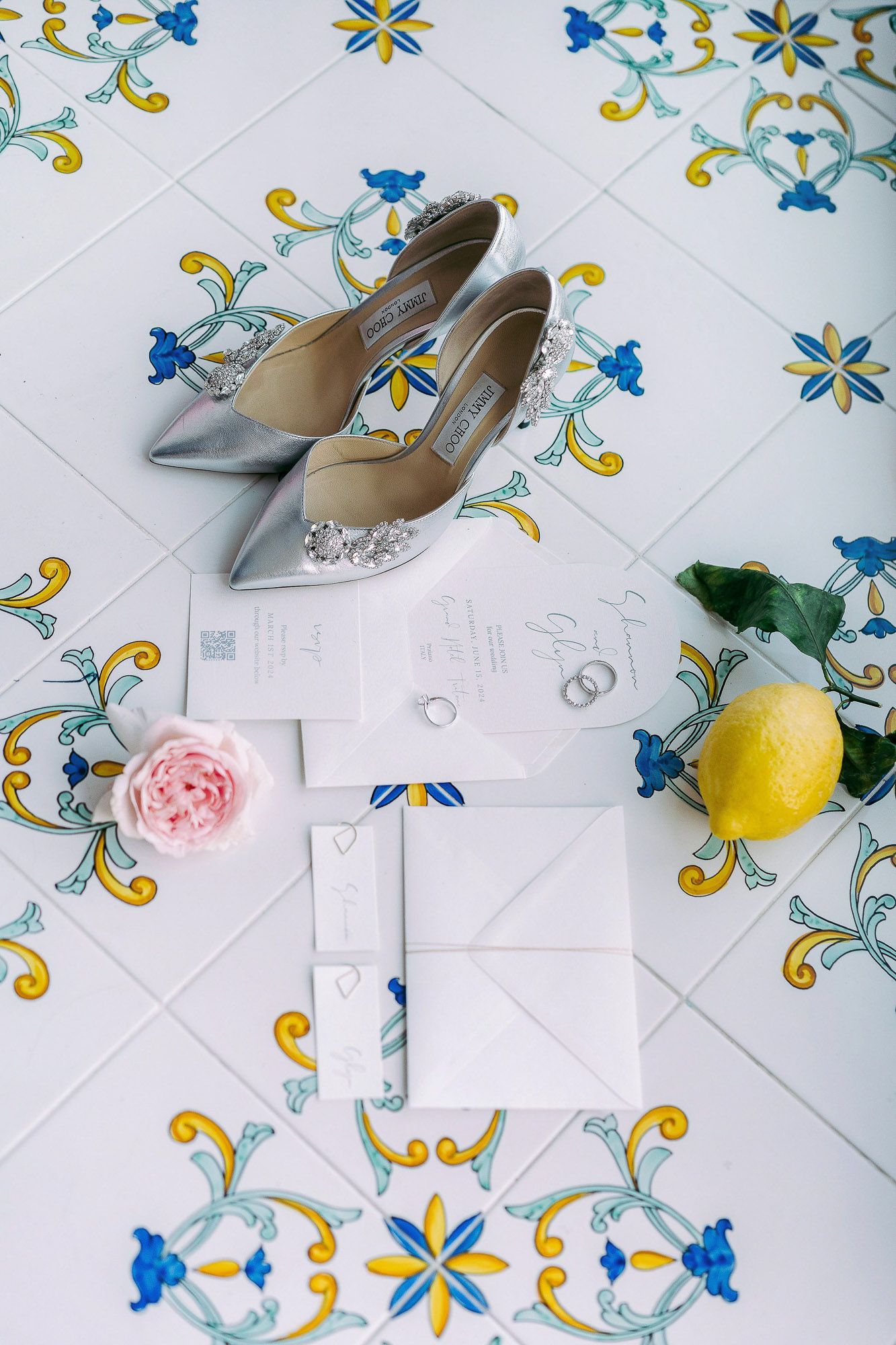 Wedding accessories including silver heels, rings, and invitations arranged on blue and yellow floral tile.