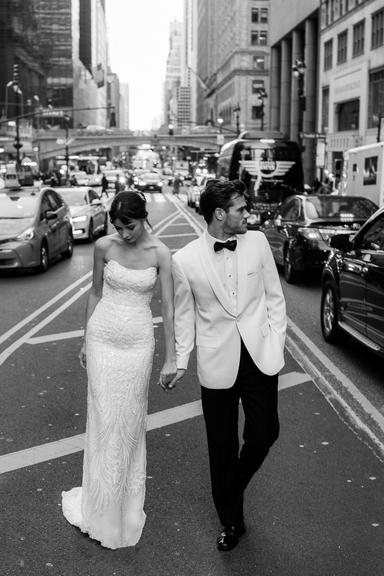 A wedding couple walks down a New York City street with tall buildings and cars in black and white.