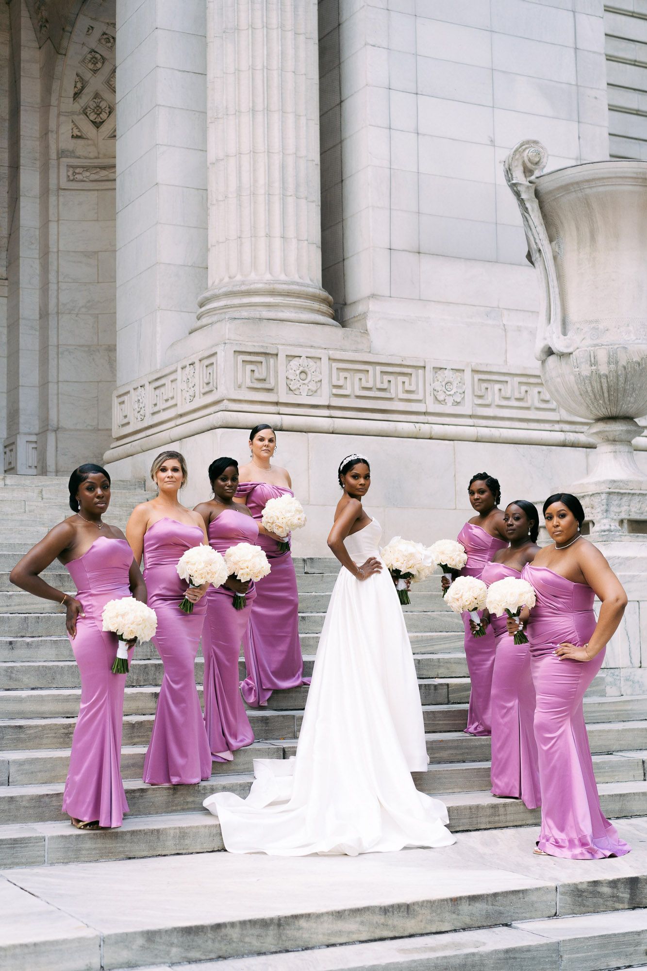 Wedding party in purple bridesmaid dresses pose on white marble steps of grand building for formal photos.