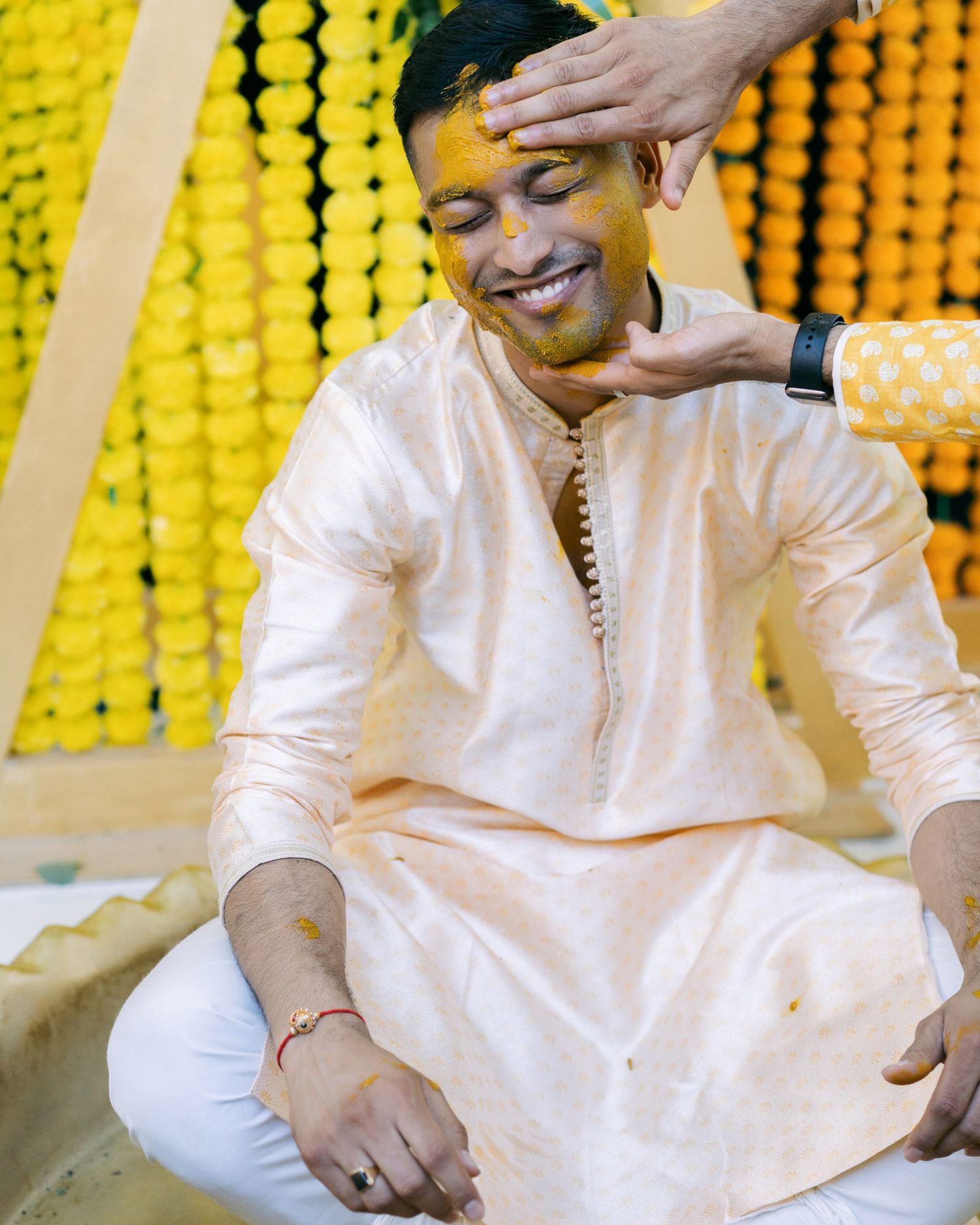 A person wearing a white traditional kurta sits and smiles during a festive celebration with yellow marigold decorations.