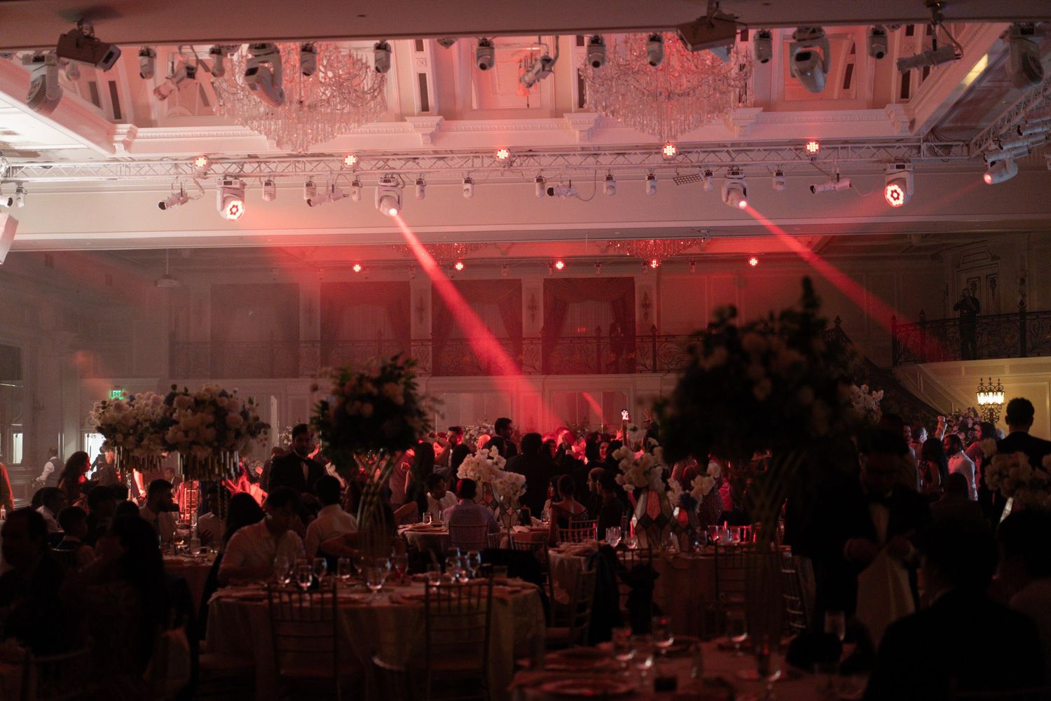 Red lighting illuminates a crowded indoor event space with guests seated at round tables during a formal gathering.