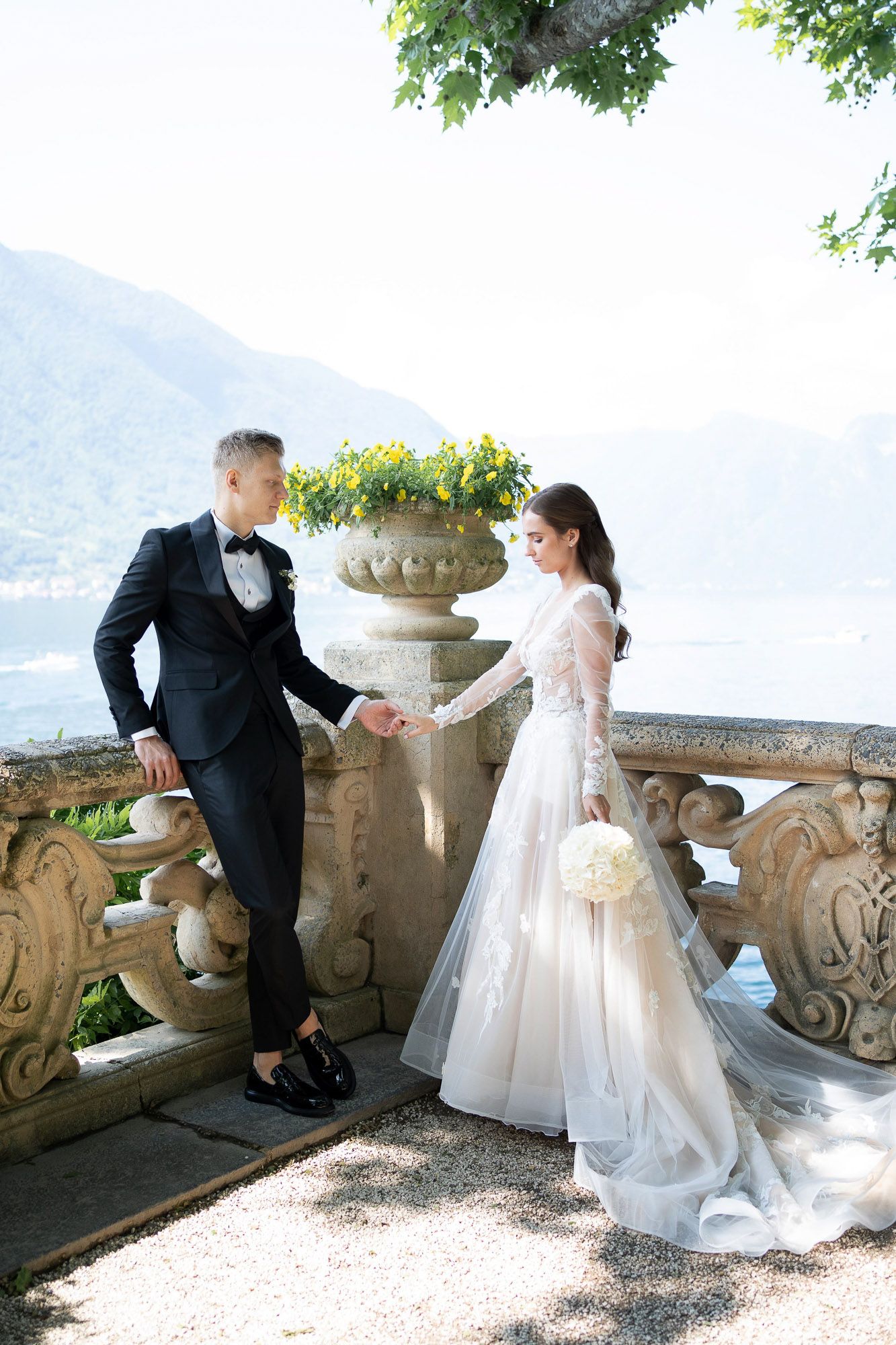 A romantic wedding couple shares tender moments on an ornate stone balcony overlooking a scenic lake view in Italy.