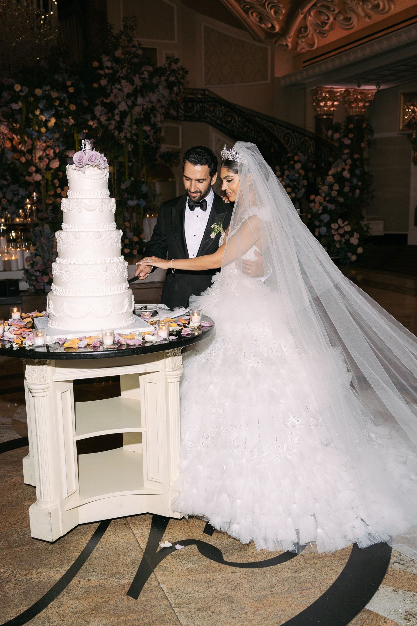 Elegant wedding couple cuts into their classic white tiered cake at luxurious indoor reception venue with floral decor.